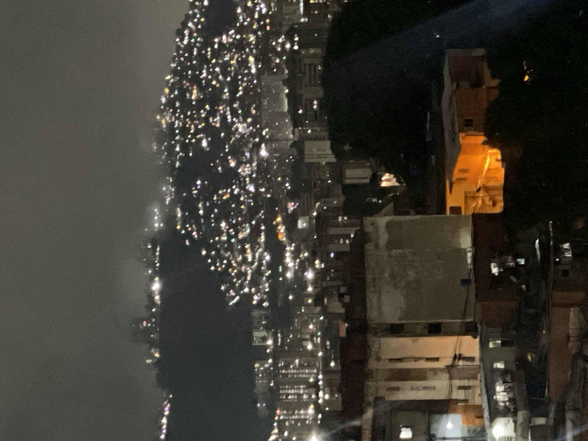 The image shows a night view of Caracas, Venezuela. A dense urban area with many buildings and streetlights is visible. A tall, multi-story structure is illuminated with a warm light, contrasting with the darker surroundings. Fog is present in the distance.