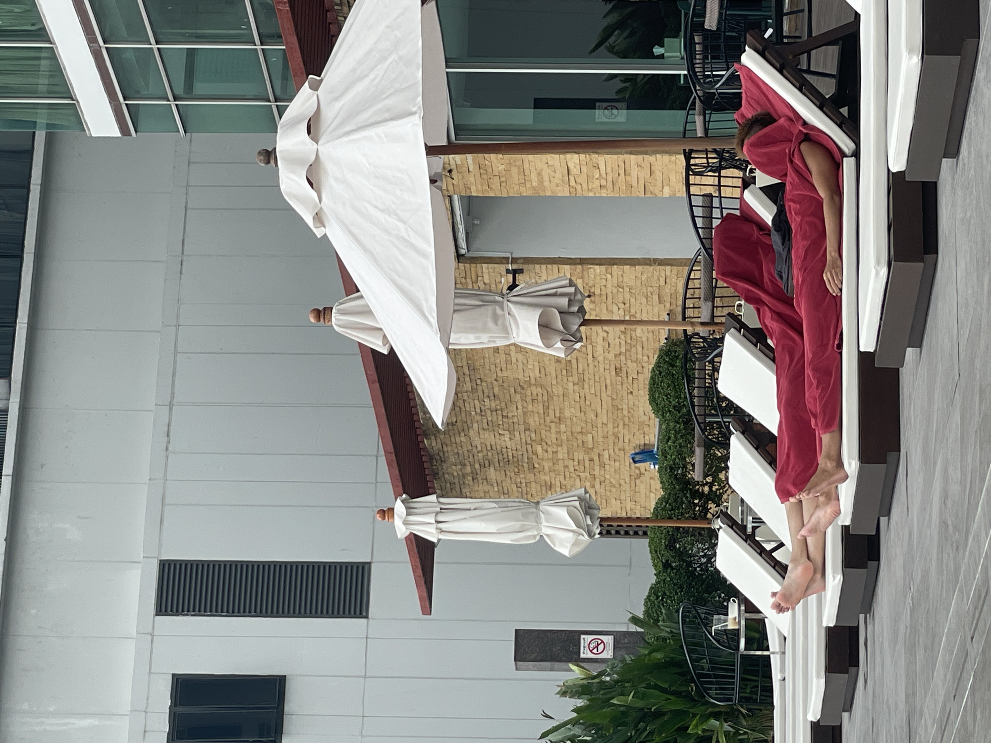 This image captures an outdoor leisure area, likely a hotel or resort, in Bangkok, Thailand. The scene features a row of white lounge chairs with dark frames, positioned on a light gray tiled surface. One person is seen lying on a lounge chair, mostly covered by a red towel or fabric, with their bare feet and lower legs visible extending towards the foreground.
Several large, white patio umbrellas are prominent. One umbrella is open and provides shade, while two others are closed and neatly tied, standing upright. In the background, a multi-textured modern building dominates. The upper part of the building displays a smooth, light gray facade with large glass windows that reflect the sky and surrounding elements. Below this, there's a section of the building with a warm, light brown brick texture. Green foliage and bushes are scattered throughout the scene, adding natural elements.
Other details include black metal outdoor furniture (tables and chairs) partially visible behind the lounge chairs and umbrellas. On the gray building, there are horizontal ventilation grates and a smaller, dark window. A "No Smoking" sign, depicted as a white circle with a red border and a crossed-out black cigarette, is visible on a dark post or wall amidst the greenery. The bright lighting suggests it is daytime, and the overall atmosphere is one of relaxation.