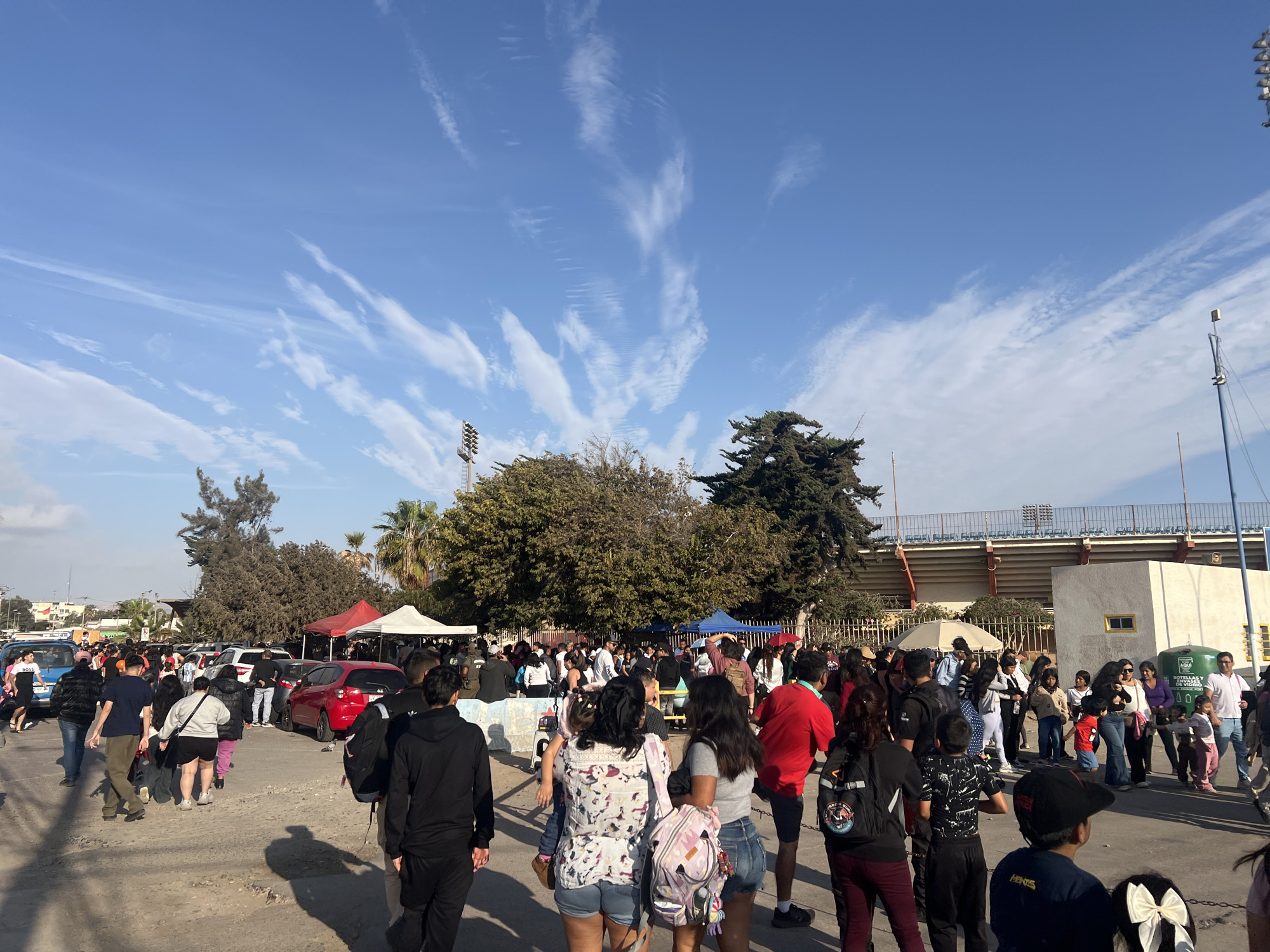 A crowd of people is gathered outdoors under a bright blue sky with scattered wispy clouds. The scene appears to be near a stadium, with stadium seating visible in the background. There are several tents set up, suggesting an event or market. Cars are parked in the area, and some individuals are walking amongst them. The ground is a rough, unpaved surface. The overall atmosphere is busy and somewhat crowded. The stadium in the background is likely the Estadio Carlos Dittborn, a prominent landmark in Arica, Chile, known for hosting sporting events and concerts.