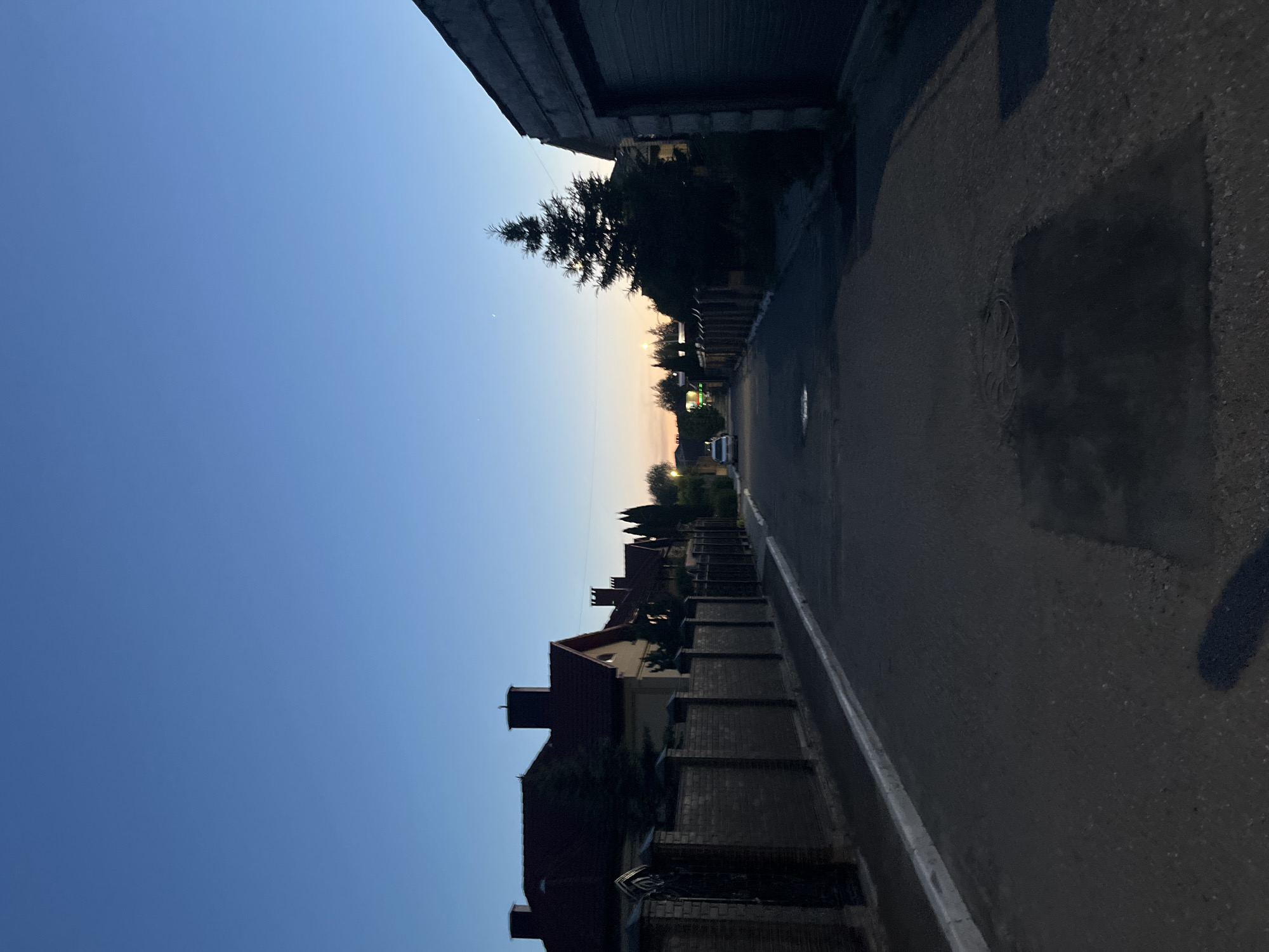 The image captures a tranquil residential street at dusk, presented as if correctly oriented with the sky at the top and the road at the bottom. The clear sky transitions from a deep, rich blue at the top to a soft, warm orange-yellow hue near the horizon, signifying either sunrise or sunset. A faint, small crescent shape, possibly the moon or a distant star, is visible in the upper-left portion of the sky.
On the left side of the street, a series of houses with dark, gabled roofs and chimneys line the properties, separated by dark brick fences. Several trees, including a prominent conifer, are interspersed along this row of homes. Further down the street, artificial lights are illuminated, and a white car is faintly visible.
The asphalt road occupies the lower and right sections of the frame. It is dark, with a distinct white line marking its left edge. Several wet patches or small puddles are visible on the road surface, and a manhole cover is discernible in the mid-ground. A dark, angular structure, possibly a building or wall, partially frames the upper-right corner of the image.
The scene is devoid of human activity, conveying a peaceful and quiet atmosphere. No visible text is present in the image. The deep shadows and specific sky coloration are key indicators of the twilight hour.