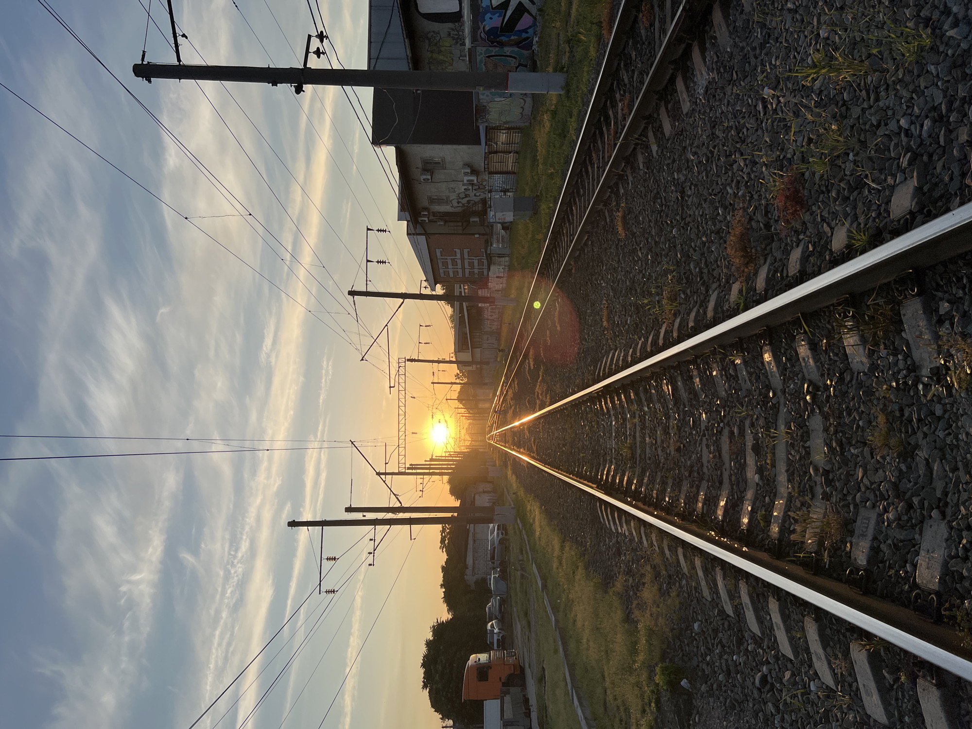 The image shows a set of train tracks stretching towards a setting sun. The tracks are surrounded by gravel and interspersed with green grass. Overhead, power lines crisscross the sky, supported by utility poles. To the left of the tracks, there is a row of parked vehicles, including an orange truck, and some buildings. To the right, industrial-looking buildings with graffiti are visible. The sky is a mix of blue and orange hues, with wispy clouds. The sun is low on the horizon, casting a warm glow and creating reflections on the metal tracks. The scene appears to be in an industrial or semi-urban area. There are no people visible in the image.
Krasnodar, Russia is a city with a significant industrial sector and railway infrastructure, so this scene is plausible within the city's landscape. The presence of industrial buildings, train tracks, and overhead power lines is consistent with areas near freight yards or industrial zones within Krasnodar. The sunset suggests it is either early morning or late evening.