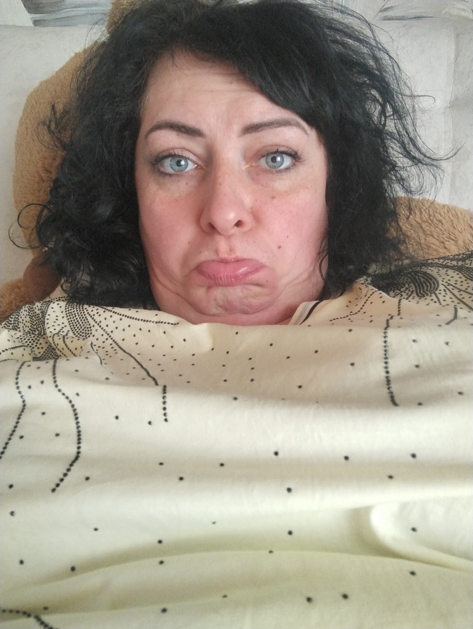 A woman with blue eyes and dark, curly hair is lying in bed. She has a neutral to slightly sad facial expression, with her lips pursed. She is covered with a light-colored patterned blanket. A brown, plush object is visible behind her head, suggesting she may be lying on a pillow or stuffed animal. The background is out of focus. The location of the photo in Grishanki, Russia, cannot be determined from the image.