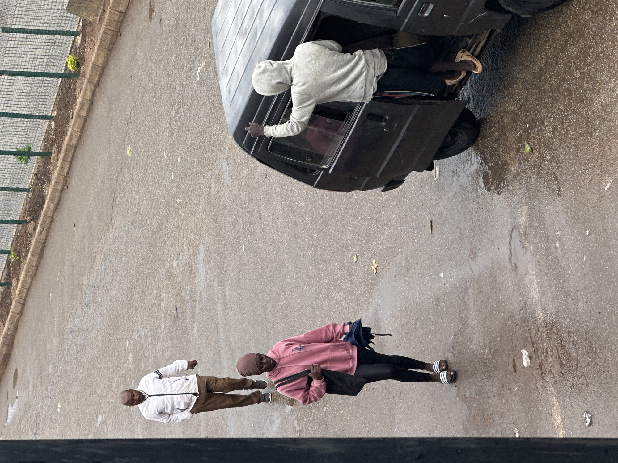 In Bukuru, Nigeria, on a wet, asphalt road, a bald Black man wearing a white hooded jacket and brown pants walks away from the camera. To his left and closer to the camera, a Black woman wearing a pink hooded sweatshirt, dark leggings, and a brown headscarf walks towards the camera, carrying a black bag and a dark umbrella. To the right of the frame, a Black person wearing a grey hooded sweatshirt and black shorts stands with their upper body leaning into the open side door of a dark-colored van, with their right arm raised as if waving or signaling. Their feet are visible in tan flip-flops. The environment suggests a cloudy day, with puddles on the road surface.