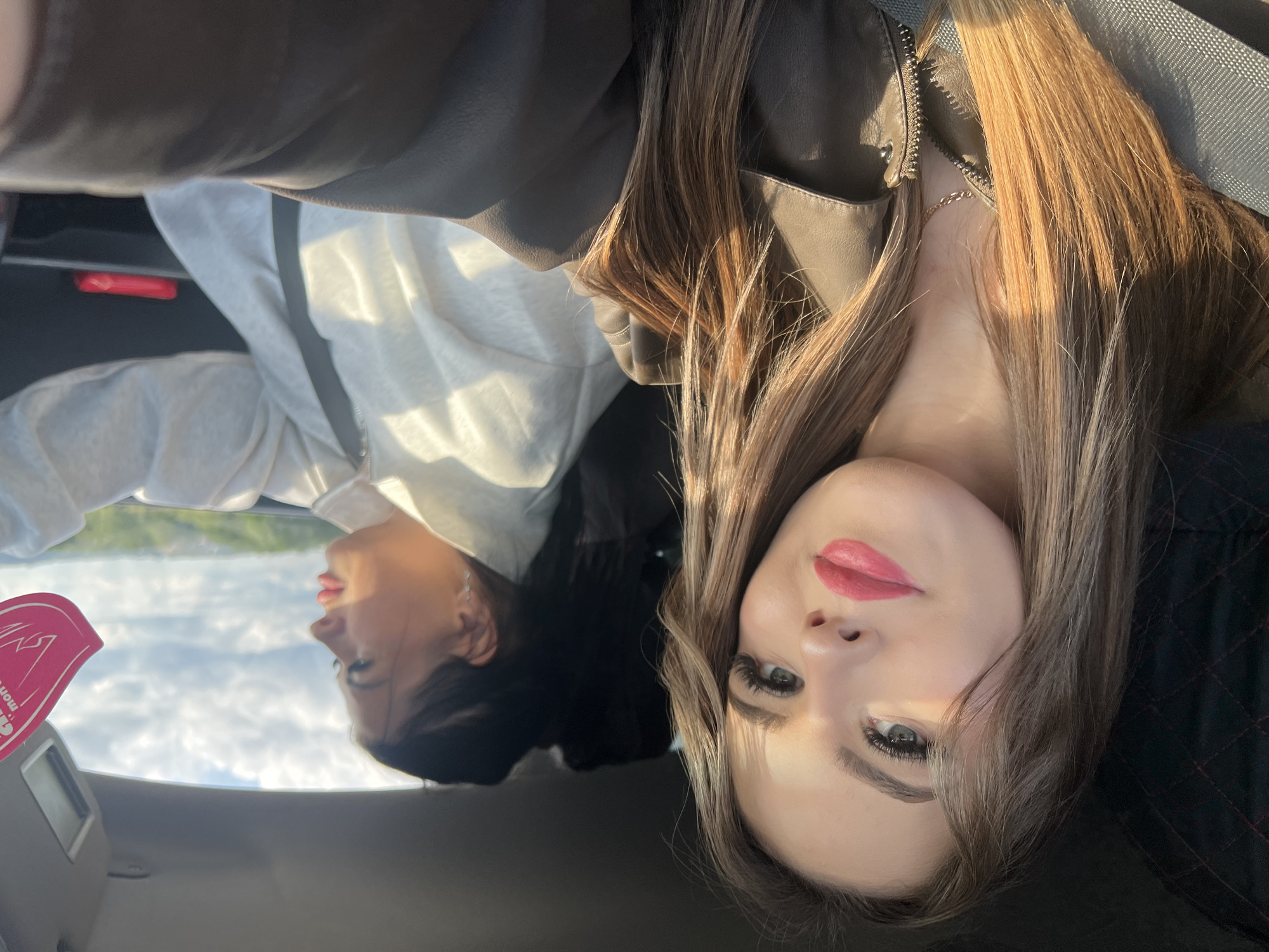 Two women are in a car. One woman has long, blonde hair and is wearing a brown leather jacket and a silver necklace. The other woman is wearing a white sweatshirt. The car is moving. Through the window is a view of land and cloudy sky. Based on the presence of a sticker written in Russian on the car's sun visor, the location is likely Russia. The city is not discernible based on the image.