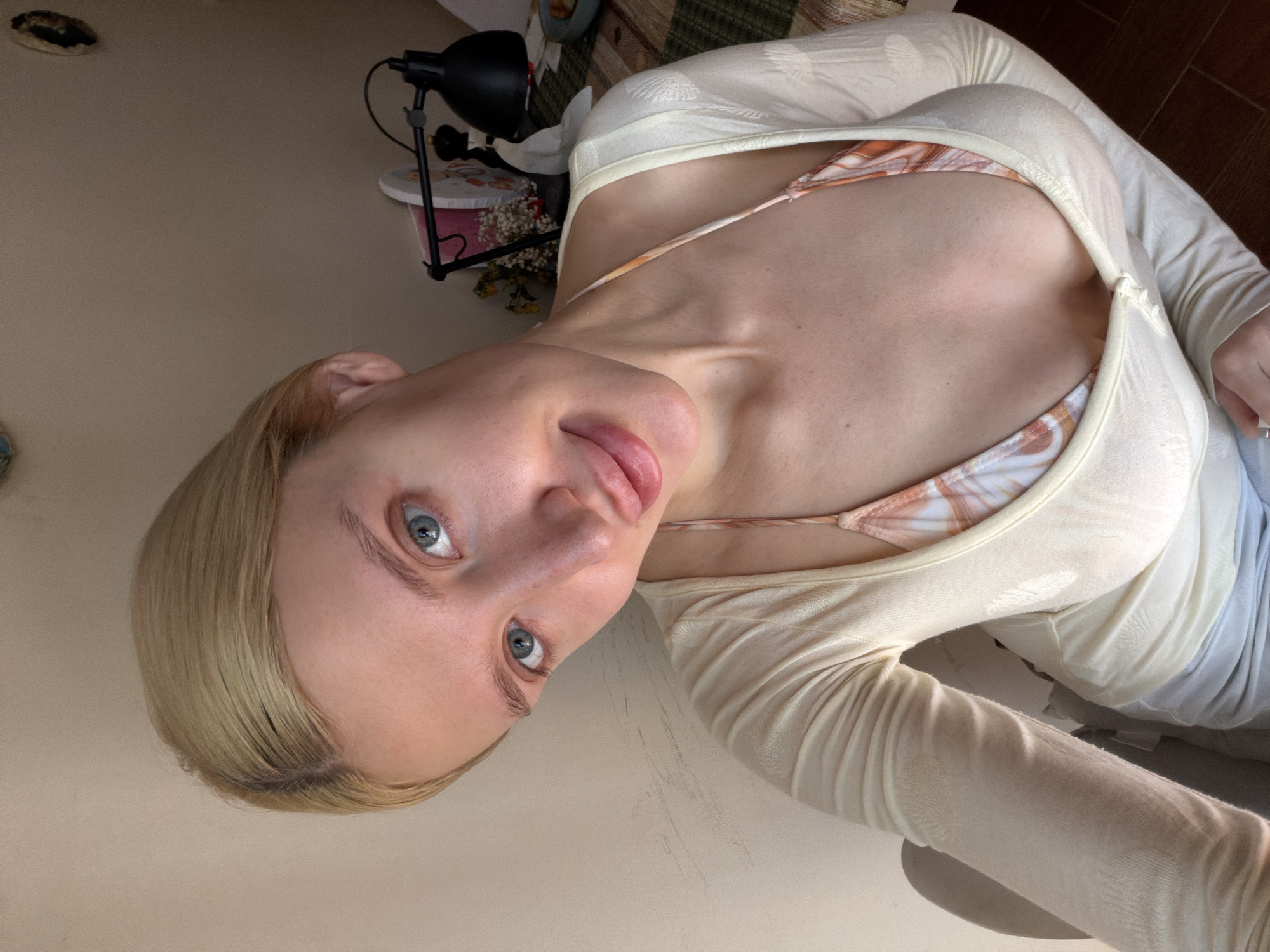 An indoor, rotated selfie captures a young woman with fair skin, blonde hair, and striking blue eyes, looking directly at the camera with a subtle, pensive expression. Her lips are slightly parted. She is wearing a light-colored, sheer, long-sleeved top over a multi-colored bra or bikini top with an orange, white, and brown pattern.
The background, in what appears to be an indoor setting in Nerin, Montenegro, features a plain light brown wall. To her side, a black desk lamp stands on a surface, beside a white container adorned with a pink pattern, holding dried flowers or potpourri. Further back, a patterned fabric, possibly a bedspread or tapestry, is visible. The scene is illuminated by natural daylight. No text is visible in the image.