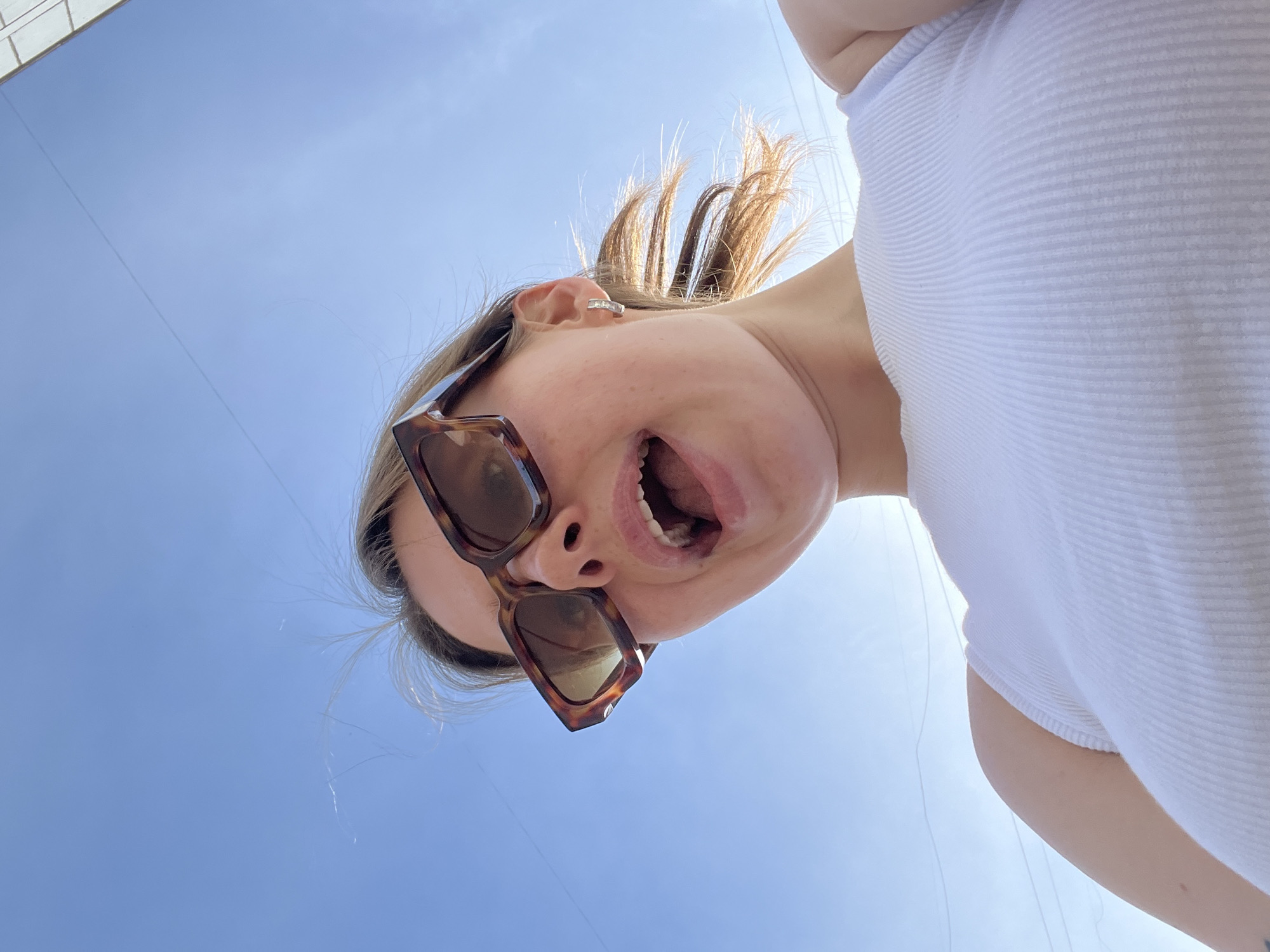 A photograph, taken from a low angle, depicts a person against a clear blue sky. The person, who appears to be a young woman, is wearing sunglasses and a white tank top. Her mouth is open, and her hair is pulled back. A building corner is faintly visible in the upper left. The location is Chaikovsky, Russia.