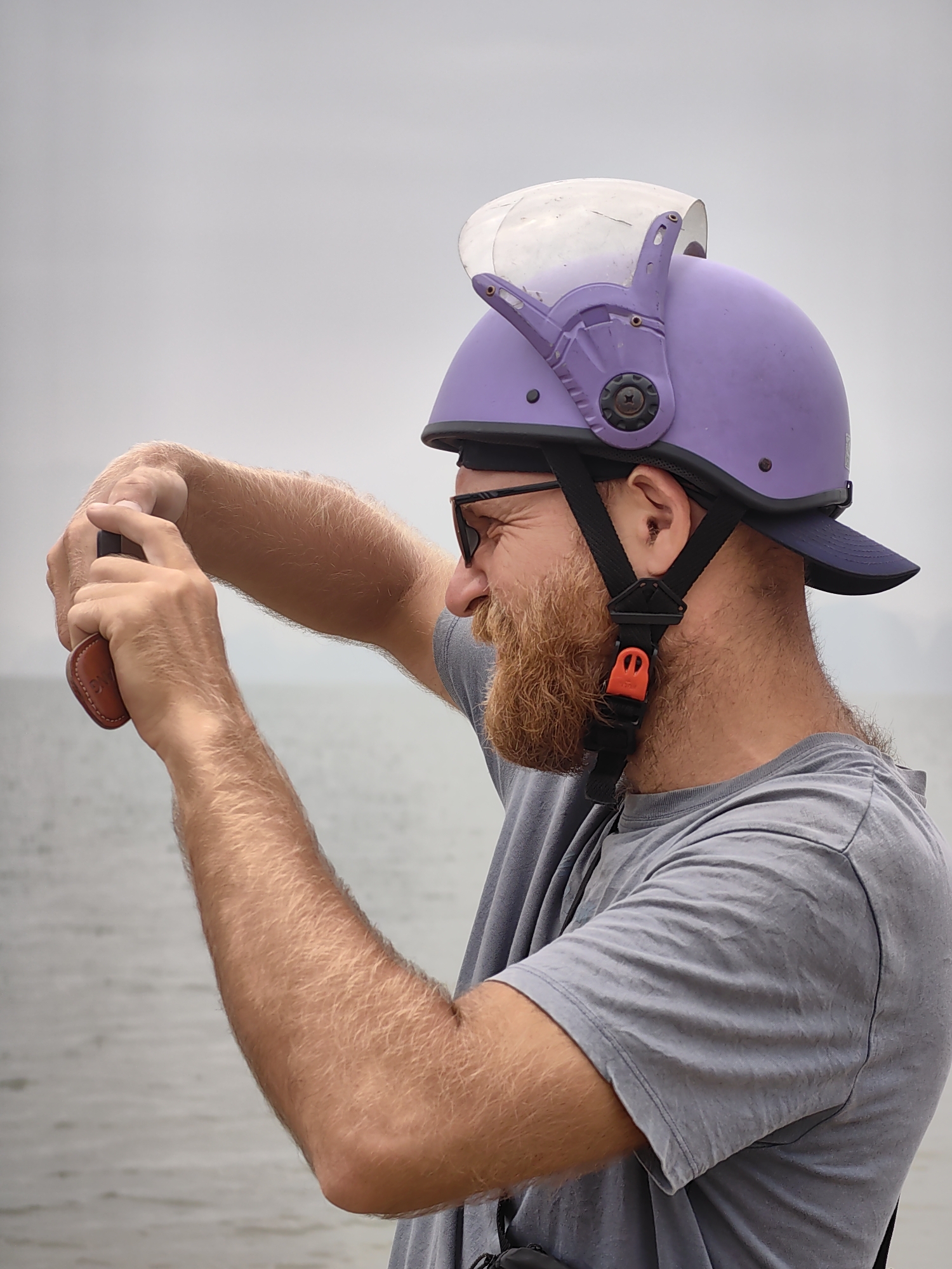 A medium shot captures a man, likely in his late 30s or early 40s, standing outdoors. He has a fair complexion, reddish-brown beard, and wears dark sunglasses. He is wearing a grey t-shirt and a unique purple helmet with a clear visor and a dark blue brim underneath. The helmet is secured by black straps with an orange buckle. The man is holding a small brown object in his hands, appearing to focus on it.
The background suggests a waterfront location. Water is visible, along with a distant landmass under an overcast sky. Considering the reference to Bai Chay Ward, Vietnam, it is plausible the photo was taken on or near the coast, perhaps overlooking Ha Long Bay.