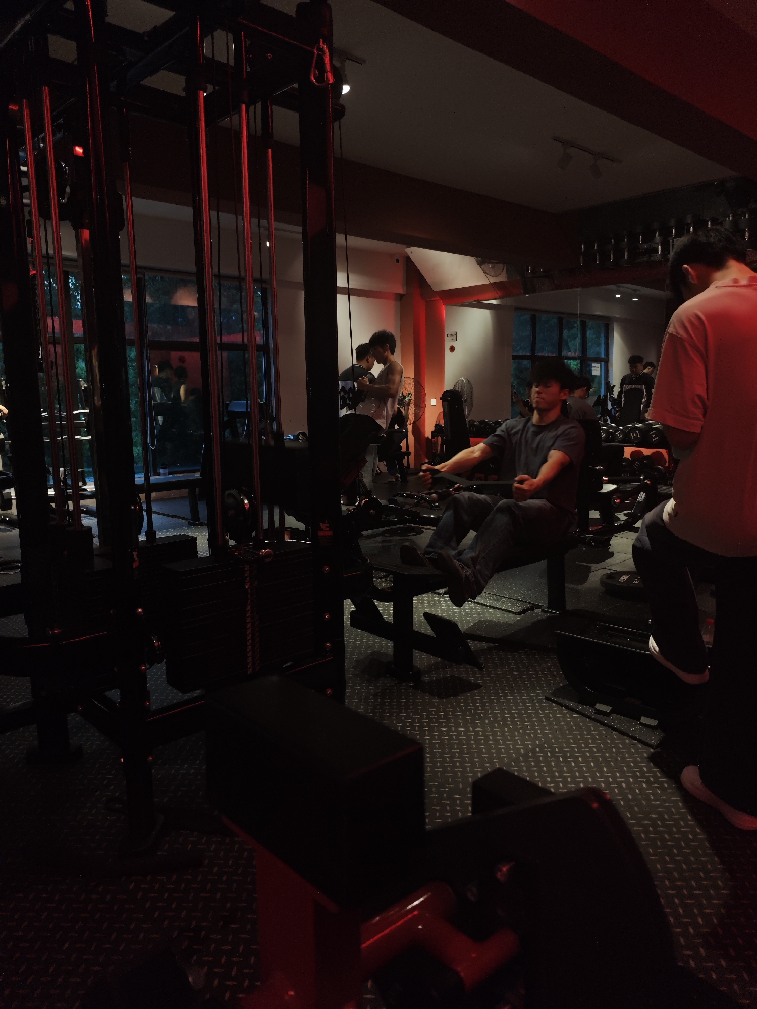 The image depicts a gym in the Chongqing High-tech Zone, China. Several people are present, including one man sitting on a rowing machine and exercising. A person is standing and wearing a red shirt is positioned in the right foreground. Other gym-goers are in the background, engaging in activities and talking to each other. The gym features various weight machines, including a cable machine with visible red cables, as well as dumbbells. The floor has a textured, possibly metallic, surface. The scene is illuminated by a mix of overhead and possibly colored lighting, creating a dim and atmospheric environment.