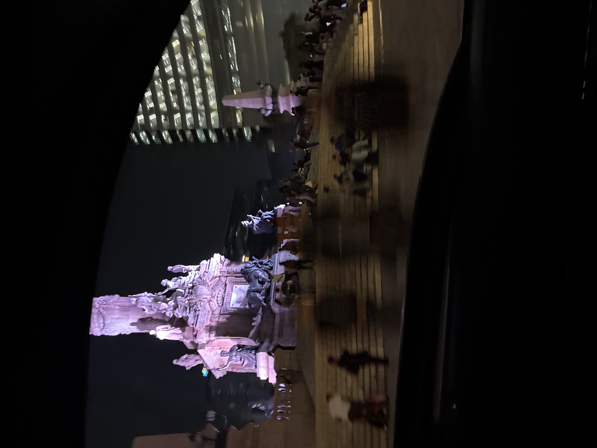 The image, taken at night, captures a vibrant urban scene featuring a large, illuminated monument and a modern high-rise building, viewed from inside a vehicle.
The central subject is an ornate, classical-style monument, dramatically lit with a purplish hue. It stands on a multi-tiered base adorned with numerous detailed sculptures, including several human-like figures in various poses. A tall, column-like structure rises from the base, topped with what appears to be another significant sculptural element, possibly a winged figure. The monumental structure strongly resembles the Monumento a la Independencia (Angel of Independence) in Mexico City, which is frequently lit up at night.
Around the monument, a crowd of people is gathered, populating the steps and surrounding area. They appear to be observing the monument, socializing, or simply enjoying the public space. Due to the low light and distance, individual faces and precise details are indistinct, but their presence suggests a lively atmosphere.
In the background, a modern skyscraper with numerous illuminated windows stands tall, providing a contrasting backdrop to the historic monument. The grid-like pattern of lights from the building's windows adds to the urban nighttime ambiance.
The photo appears to have been taken from a slightly elevated position, possibly through a car window, as indicated by the dark, curved elements framing the bottom and left edges of the frame. There is some slight motion blur, particularly among the people and in the building lights, suggesting either a handheld shot in low light or movement of the camera or vehicle. No discernible text is visible in the image.