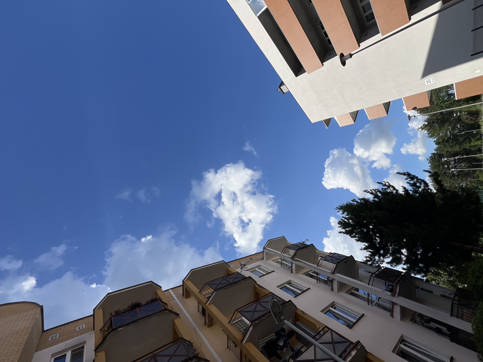 This is a low-angle, upward-facing photograph of apartment buildings against a bright blue sky with scattered white clouds. The scene captures the architecture of two distinct buildings.
The building on the right is taller and modern, with a light-colored facade and prominent horizontal bands of a reddish-brown material. Its balconies are recessed, and some have dark railings. A sign with the text "2G" is visible on its side. To the right of this building, the tops of tall evergreen trees suggest a natural setting.
The building on the left appears older, constructed with light brown brick on its upper sections and a lighter colored lower section with balconies. These balconies are more prominent, with dark railings and a decorative metal grid pattern. Some balconies have small planters with flowering plants.
The image seems to be taken on a clear, sunny day, possibly in the late morning or early afternoon, given the bright sky and distinct shadows. The overall impression is of a residential area in an urban or suburban environment, likely in Poland, as indicated by the location context. No people are visible in the image. The low-angle perspective emphasizes the scale of the buildings and the expanse of the sky.