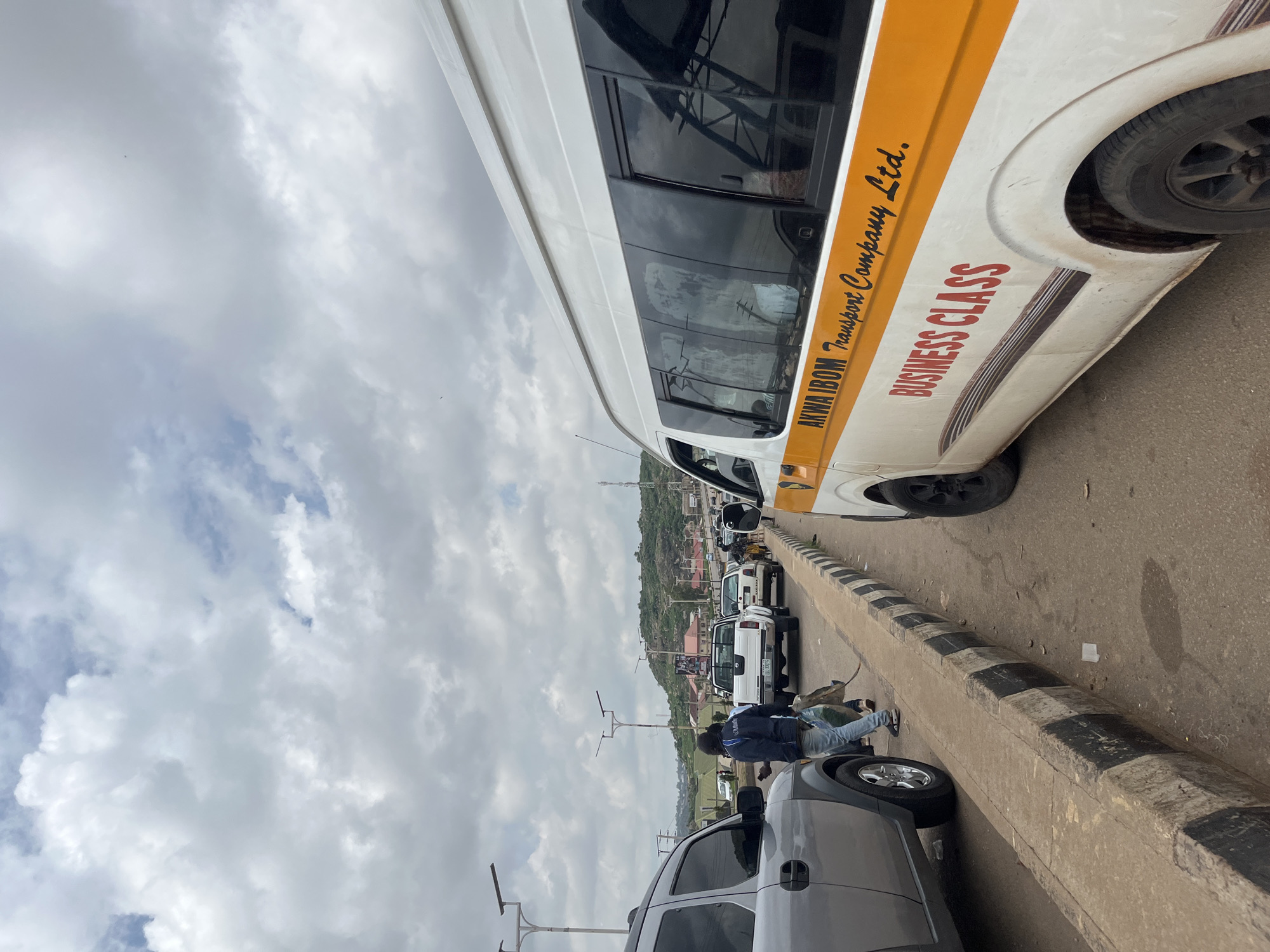 The image depicts a street scene in Gura Top, Nigeria. A white van with orange and black decals is prominently featured on the right side of the frame, displaying the text "AKWA IBOM Transport Company Ltd." and "BUSINESS CLASS" in red lettering. The van appears to be in motion or stopped in traffic. To the left, a silver SUV is partially visible, also seemingly in traffic. Several other vehicles, including white pickup trucks and cars, are observed on the road. A pedestrian, wearing a dark jacket and blue jeans, is walking on the sidewalk carrying a large bag. The environment is urban, with paved roads, a concrete curb marked with black and white stripes, and overhead streetlights. In the background, buildings and a hilly landscape are visible under a cloudy sky. The overall impression is of a busy, possibly congested, street in Nigeria.