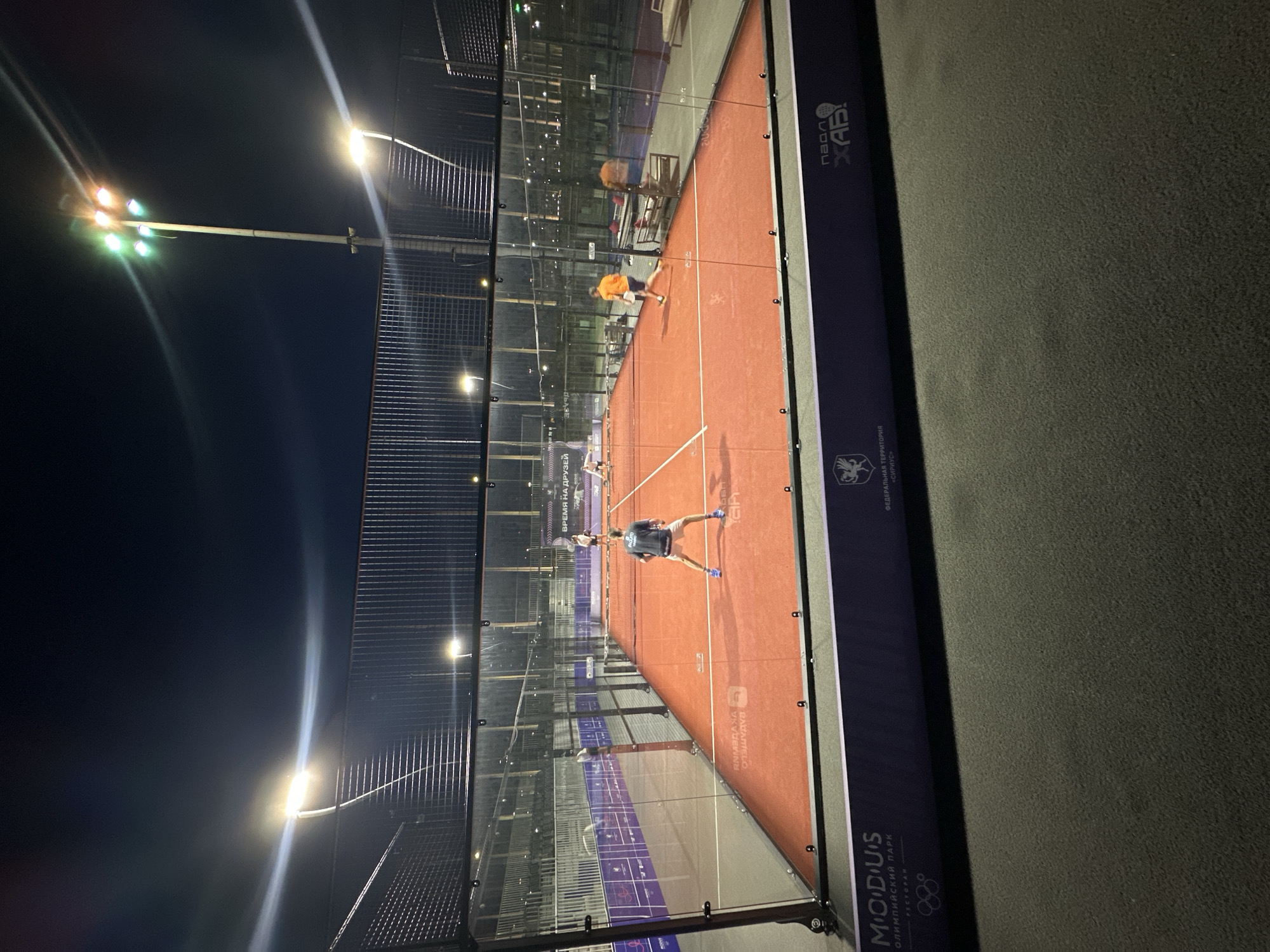 The image captures a padel tennis match taking place at night on a brightly illuminated court within the Federal Territory "Sirius," Russia. The court's playing surface is a distinct bright orange-red, marked with white lines, and enclosed by clear glass walls topped with black wire mesh fencing.
Four male players are actively engaged in the game. In the immediate foreground, facing away from the camera, a player in a dark grey t-shirt, light shorts, and blue athletic shoes holds a padel racket, positioned near the service line. Opposite him, across the black net, a player in a white t-shirt and dark shorts stands near the net. To the right, another player in a vibrant orange t-shirt and dark shorts is in motion, running towards the right side of the court with a racket. A fourth player, also in a white t-shirt and dark shorts, is visible further back on the court near the rear glass wall.
The court is intensely lit by multiple floodlight poles. A prominent tall pole on the mid-right side features several bright lights, some appearing white-yellow, others with faint green and orange hues, likely due to lens flare. Additional intense white-yellow lights on shorter poles around the perimeter cast strong illumination and create significant light streaks across the dark night sky.
Behind the court's glass enclosure, other dimly lit padel courts with similar orange surfaces and spectator seating areas are discernible. Benches or resting areas are visible on both sides of the court, outside the playing area.
The base of the court structure features purple banners with white text. On the left, a banner reads "M O D U S", "ОЛИМПИЙСКИЙ ПАРК" (Olympic Park), and "РЕСТОРАН" (Restaurant), accompanied by the Olympic rings logo. A central banner explicitly states "ФЕДЕРАЛЬНАЯ ТЕРРИТОРИЯ" (Federal Territory) and "«СИРИУС»". On the far right, another banner displays "ПАДЕЛ ХАБ!" (Padel Hub!). A banner on the rear wall of the court prominently features the phrase "ВРЕМЯ НА ДРУЗЬЯ" (Time for Friends). The ground in the immediate foreground consists of a dark grey, textured surface.