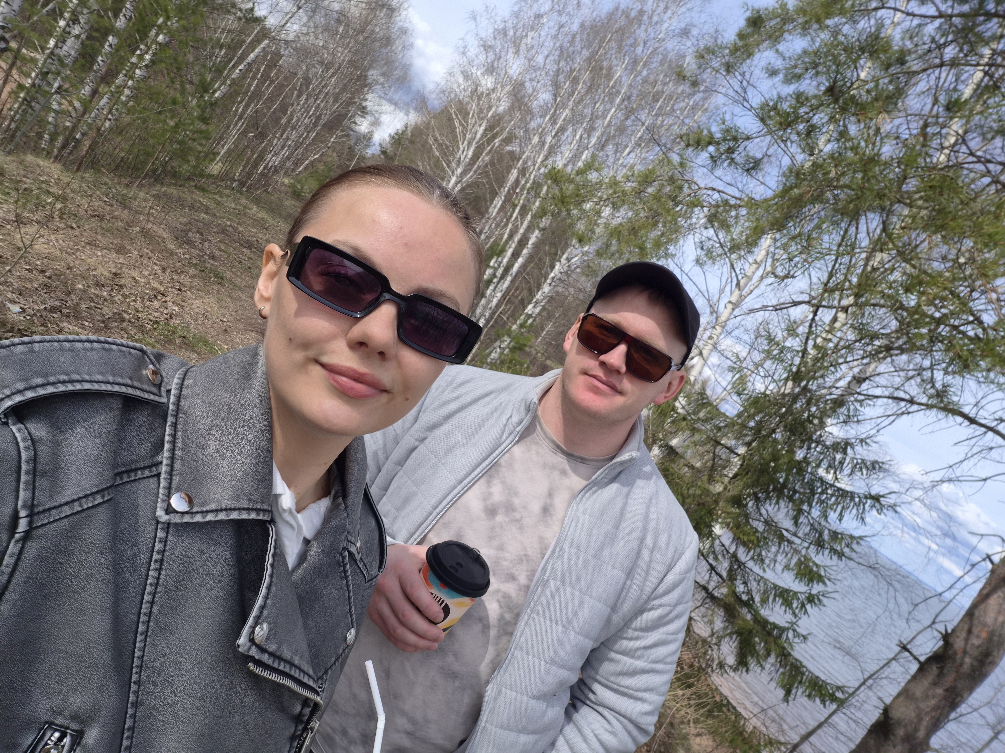 A casual selfie captures a woman and a man enjoying an outdoor day in what appears to be an early spring setting in Chaikovsky, Russia. The woman, positioned closer to the camera on the left, wears a grey faux-leather biker jacket over a white shirt, complemented by dark, rectangular sunglasses. Her hair is pulled back, and she offers a slight smile. Beside and slightly behind her, the man wears a light grey quilted jacket over a patterned light t-shirt, a dark baseball cap, and dark sunglasses. He holds a colorful disposable coffee cup with a black lid in his right hand. A white straw is visible near the bottom left, suggesting another beverage.
The background reveals a natural landscape consistent with a park or forest area. Dominating the scene are numerous birch trees with their distinctive white bark, some still bare of leaves, alongside evergreens. The ground is sparsely covered with dry leaves and patchy vegetation, indicative of early spring. To the right, a body of water, likely the Kama River or a lake, reflects the bright sky. The weather appears sunny with a few scattered clouds, providing ample natural light for the daytime scene. The overall mood conveyed is relaxed and pleasant, as the couple enjoys their outing in the natural surroundings. No clearly legible text is visible, but the coffee cup features an abstract, multi-colored pattern.