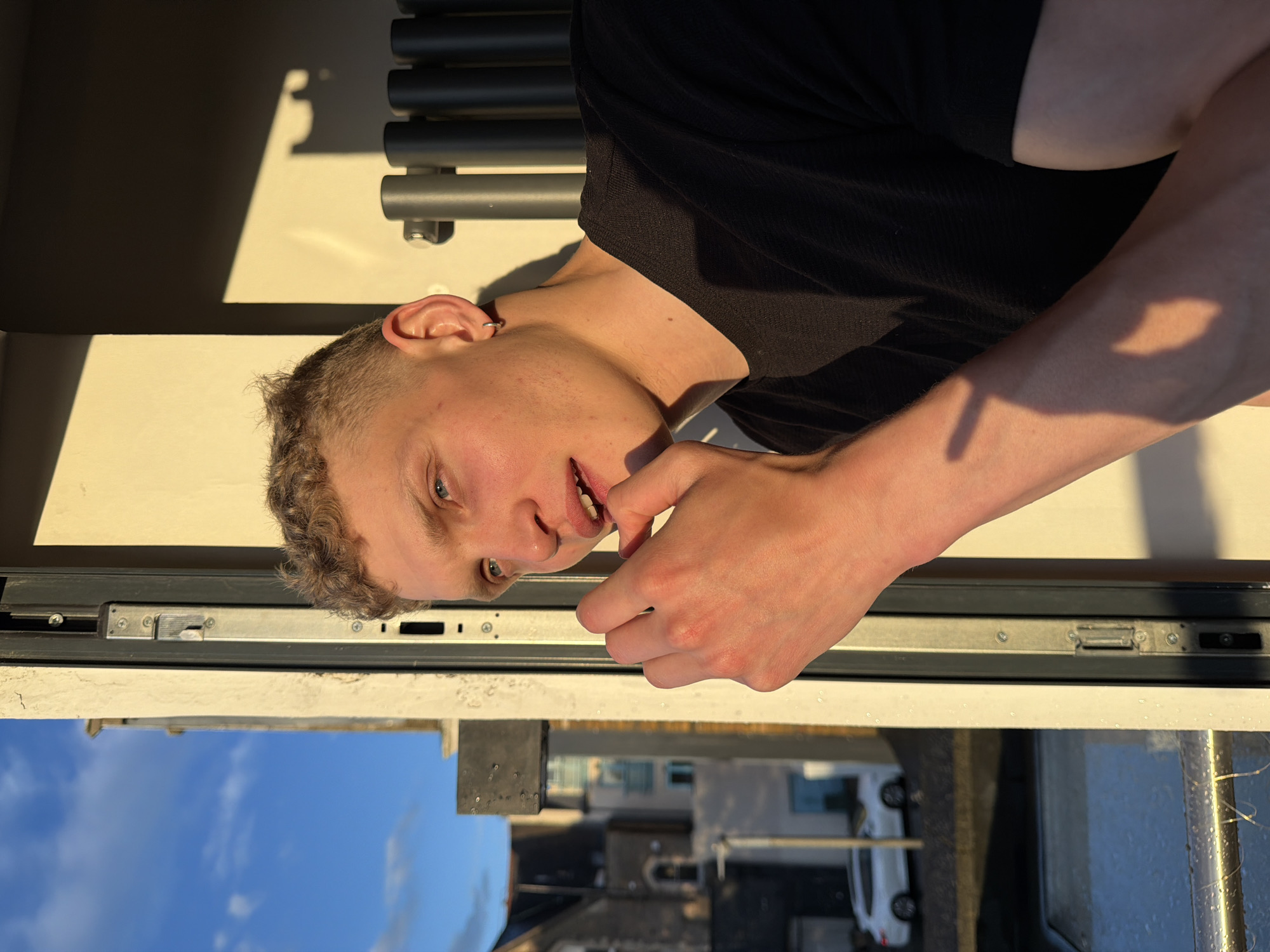 A young, light-skinned man with short curly hair and blue eyes is looking directly at the camera while seemingly biting his fingernails. He is wearing a black t-shirt. The man is standing inside near a window. Outside the window, buildings and cars are visible, suggesting an urban area. The sky is blue. The scene appears to be lit by natural sunlight. The location is unclear and cannot be definitively identified as London, United Kingdom based solely on the provided image.