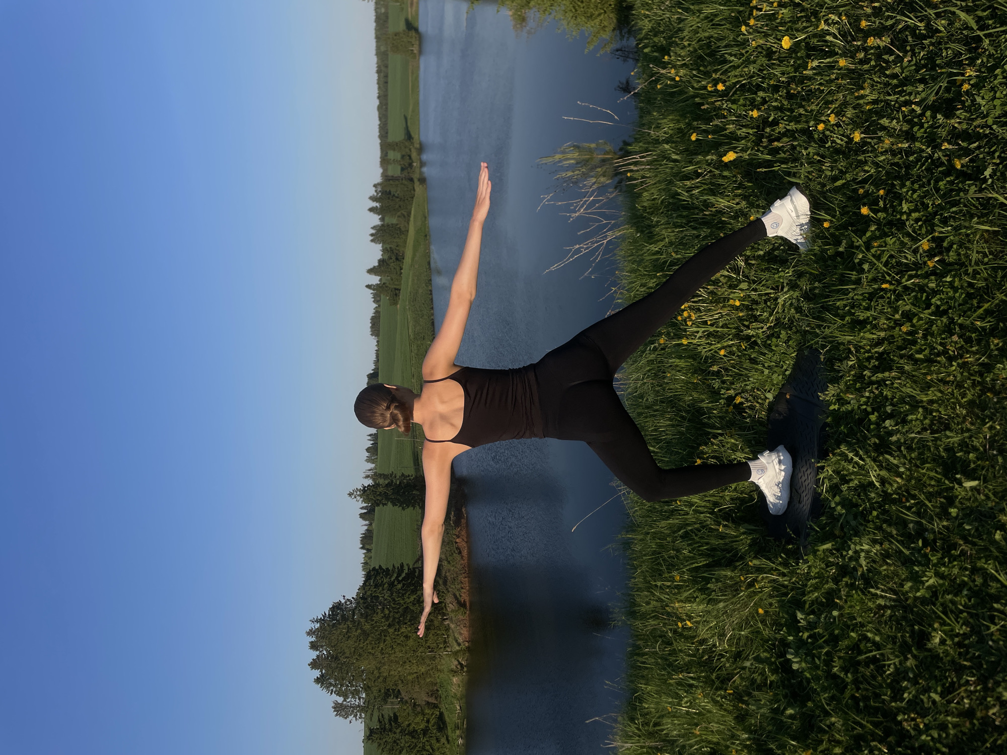 A woman is captured from behind, performing a yoga pose on a grassy terrain near a body of water. The location is Подшивалово, Russia. She is clad in black athletic attire, including a tank top and leggings, and white sneakers. Her arms are outstretched, forming a line with her body. The grassy ground is dotted with yellow wildflowers, and a yoga mat is visible beneath her feet. The body of water is flanked by green fields and trees. Above, the sky transitions from a clear blue to a lighter shade near the horizon.