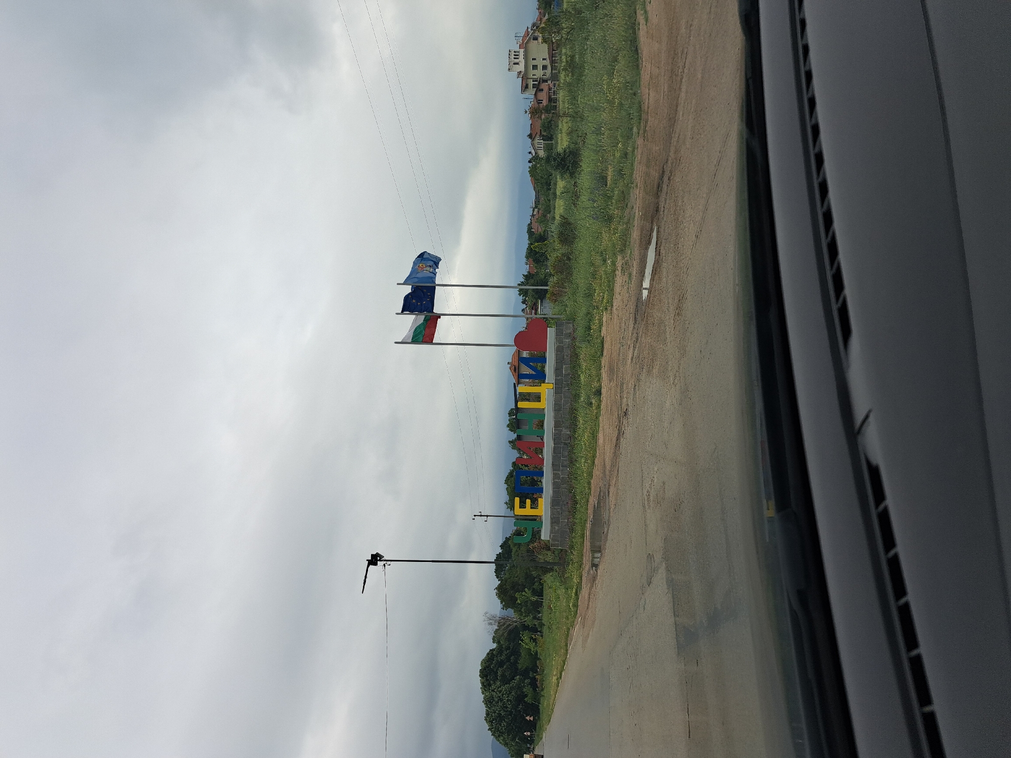 The image captures a roadside view in Dolni Bogrov, Bulgaria, on a cloudy day. A large, colorful sign spelling out "ЧЕПИНЦИ" (Chepinsi) in Cyrillic letters stands prominently on the right side of the road, constructed from individual, brightly colored blocks (yellow, blue, red, yellow, blue, red, yellow). Adjacent to the sign, three flags are flown on tall poles: the Bulgarian flag (white, green, red), the European Union flag, and another flag, possibly a regional or municipal one, in blue with a yellow emblem.
In the background, the scene features residential buildings with reddish-tiled roofs, a cluster of green trees, and a distant view of hills or mountains under the overcast sky. The foreground shows a paved road with some cracks and puddles, suggesting recent rain. A portion of a car's dashboard and windshield is visible at the very bottom of the image, indicating the photo was taken from inside a vehicle. A street lamp is positioned behind the sign, and utility poles with wires run across the scene. The overall environment appears to be a semi-rural or suburban area on the outskirts of a town.