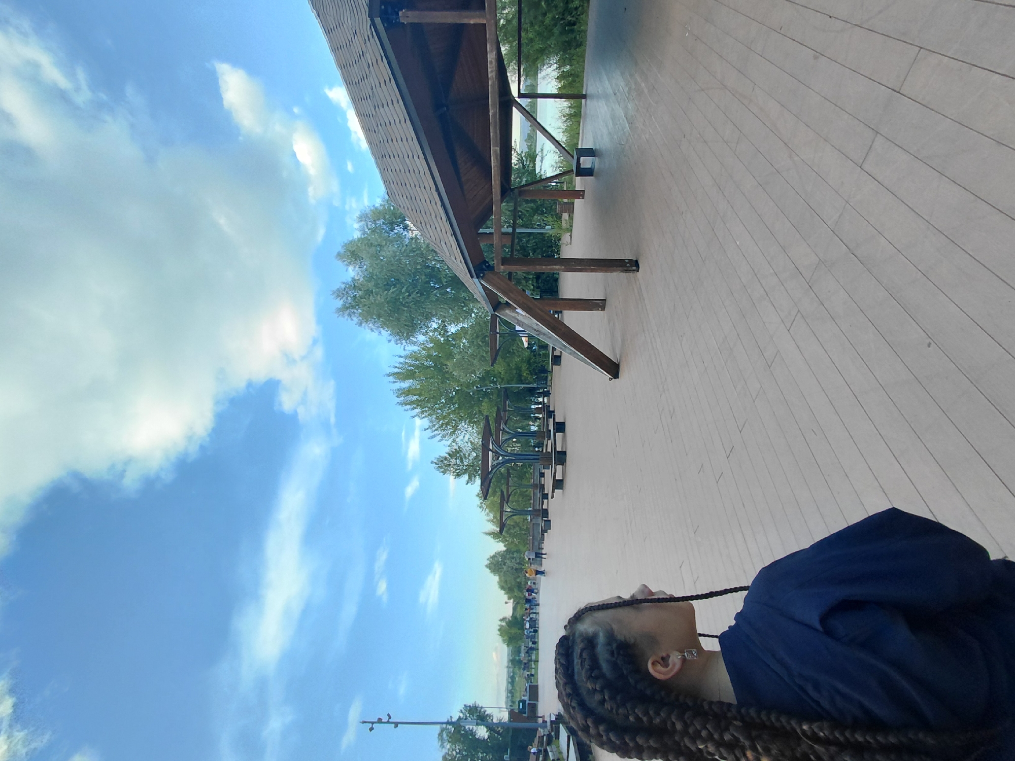 A person with braided hair and a navy blue garment is captured in a tilted selfie, looking up at the sky. The setting appears to be an outdoor wooden boardwalk, possibly part of a park or waterfront area, given the presence of trees and a distant body of water. A large, covered wooden structure, resembling a gazebo or picnic shelter, is visible in the mid-ground. The sky is a vibrant blue with scattered white clouds, suggesting a pleasant day, likely daytime. The wooden planks of the boardwalk stretch into the distance, where several picnic tables and other park amenities can be seen. A streetlamp or light pole is also visible in the background. No visible text is present in the image.
