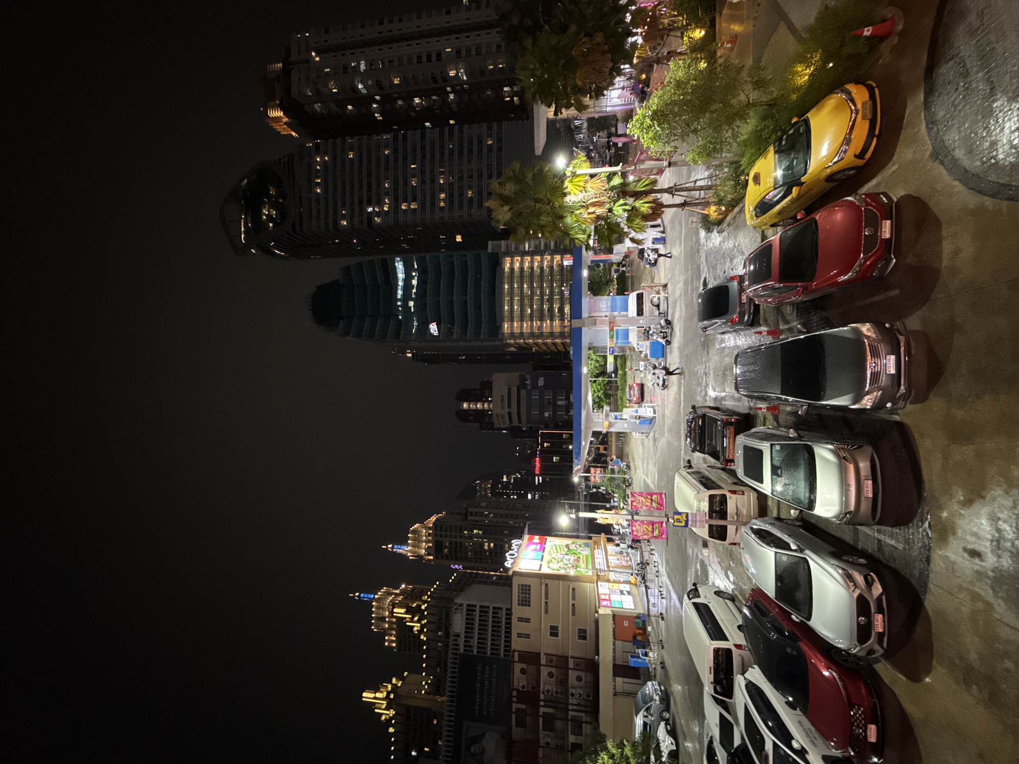 The image depicts a nighttime urban scene in Bangkok, Thailand. Tall buildings with illuminated windows dominate the left side of the frame, towering over smaller buildings and structures. On the right, a street or parking area is visible, lined with parked cars of various colors and models. Streetlights illuminate the scene, casting reflections on the wet pavement. A gas station is visible between the buildings and parked cars, with gas pumps and people moving around. The overall atmosphere suggests a busy urban environment at night.