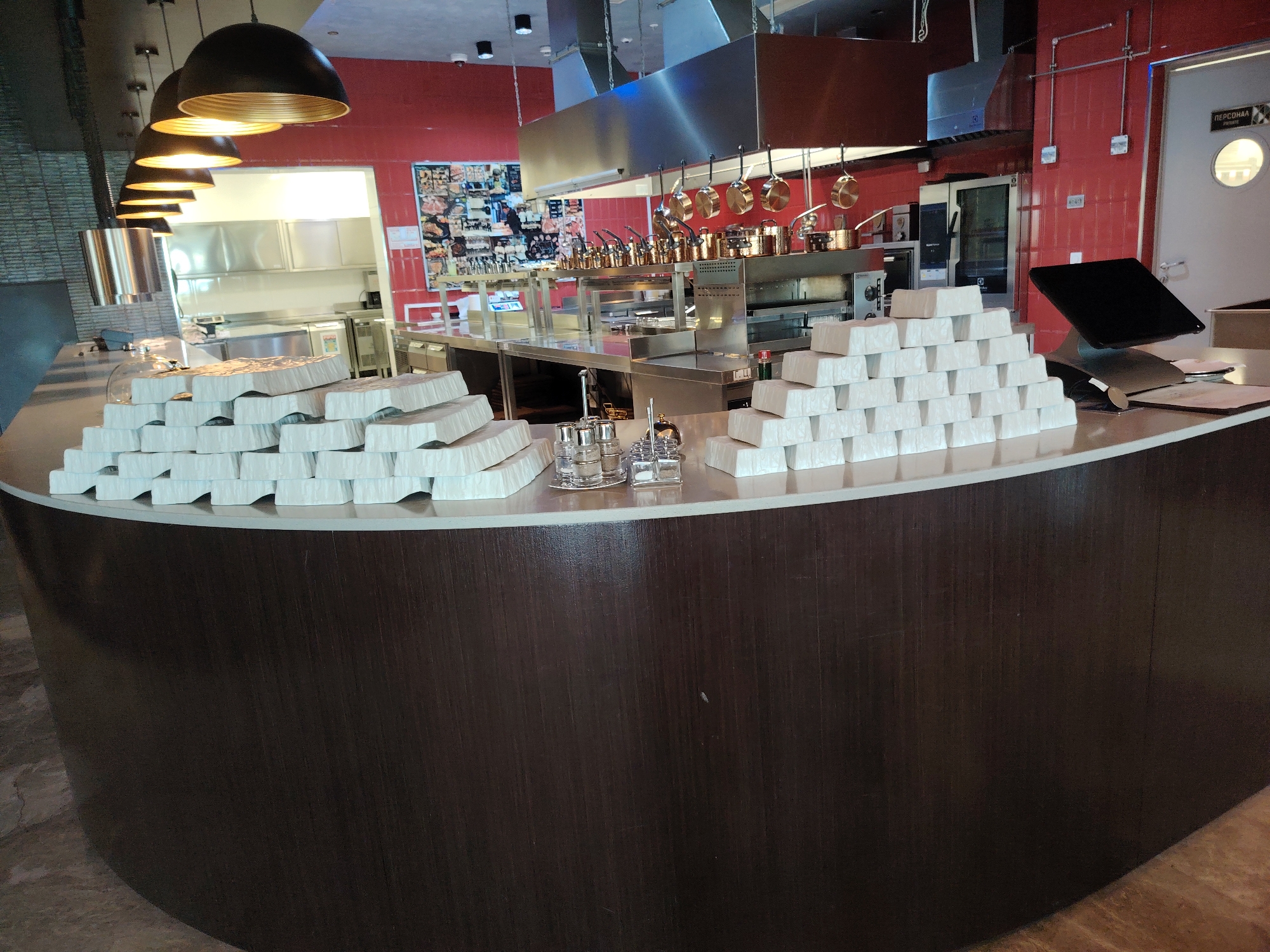 The image shows a commercial kitchen with a curved counter in the foreground. Piled on the counter are stacks of white, rectangular containers, possibly for food. In the background, stainless steel cooking equipment, including stoves and ventilation hoods, is visible. Various pots and pans are hanging above the cooking area. A door labeled "ПЕРСОНАЛ" (Personnel) is to the right, with a circular window. The walls are painted red. The floor appears to be tiled. The overall scene suggests a restaurant or catering establishment that is either preparing for service or has recently concluded a busy period.