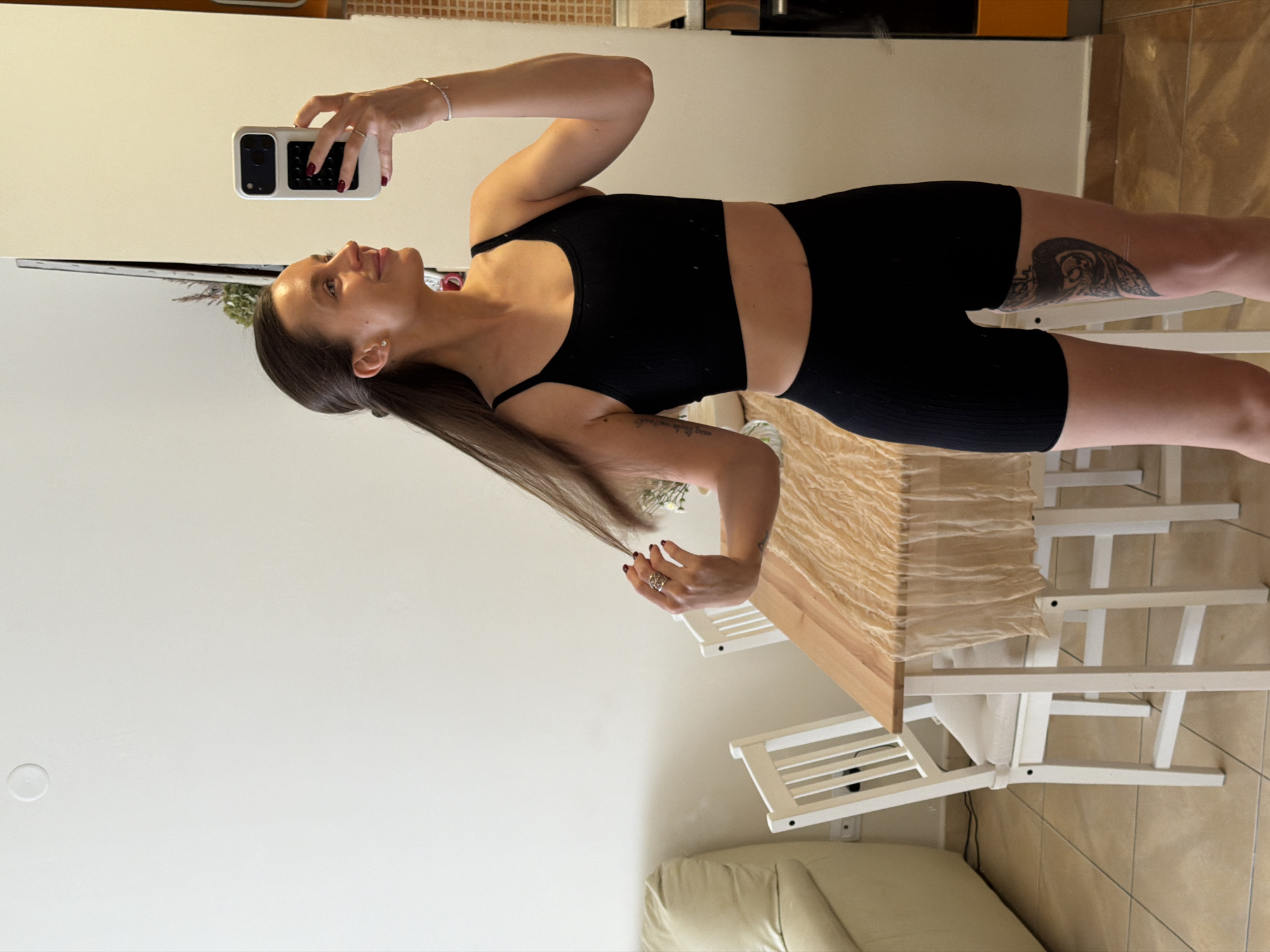 A person is taking a selfie in an indoor setting. The individual, presumably female, is wearing a black sports bra and black shorts. She is holding a smartphone in her right hand to capture the image. The background includes a white wall, a dining table with a light-colored tablecloth, and white chairs. A glimpse of a light-colored sofa is visible in the lower part of the image. There is no immediate visual indication of Budva, Montenegro. The person's left leg is adorned with a tattoo.