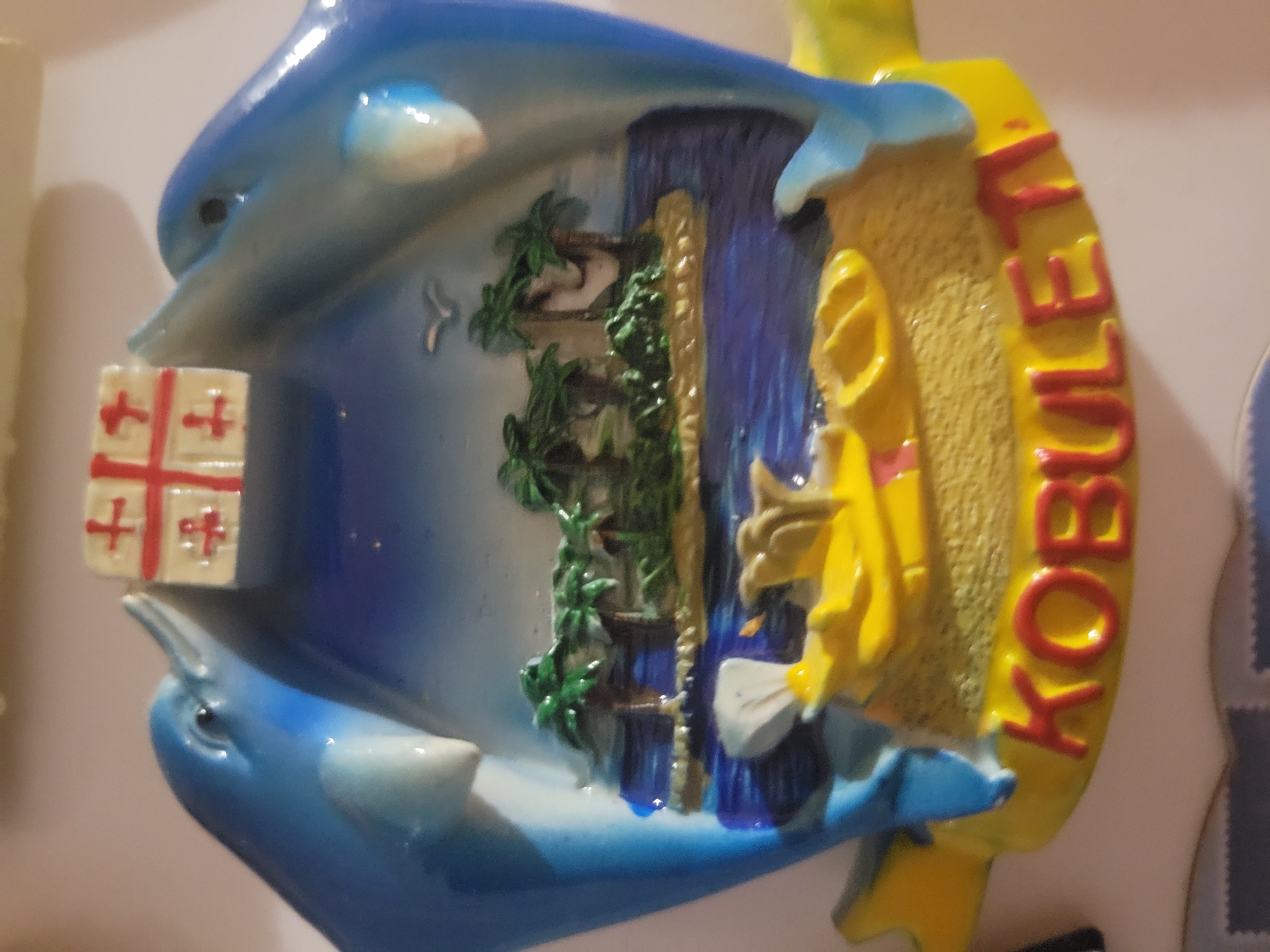 This is a close-up, eye-level, medium shot of a novelty magnet, likely a refrigerator magnet. The magnet depicts two stylized blue dolphins facing each other, with their heads nearly touching at the top. Between their heads, a small, square emblem with a red cross on a white background is visible, resembling a flag. The background behind the dolphins transitions from a dark blue, starry night sky to a tropical island scene. The island features palm trees and a sandy beach, with blue water surrounding it. In the foreground, a yellow object, possibly a stylized boat or raft, is depicted with red lettering that spells out "KOBULETI". The magnet is placed against a neutral, out-of-focus background. There is no indication of the location being Belaazyorsk, Belarus.