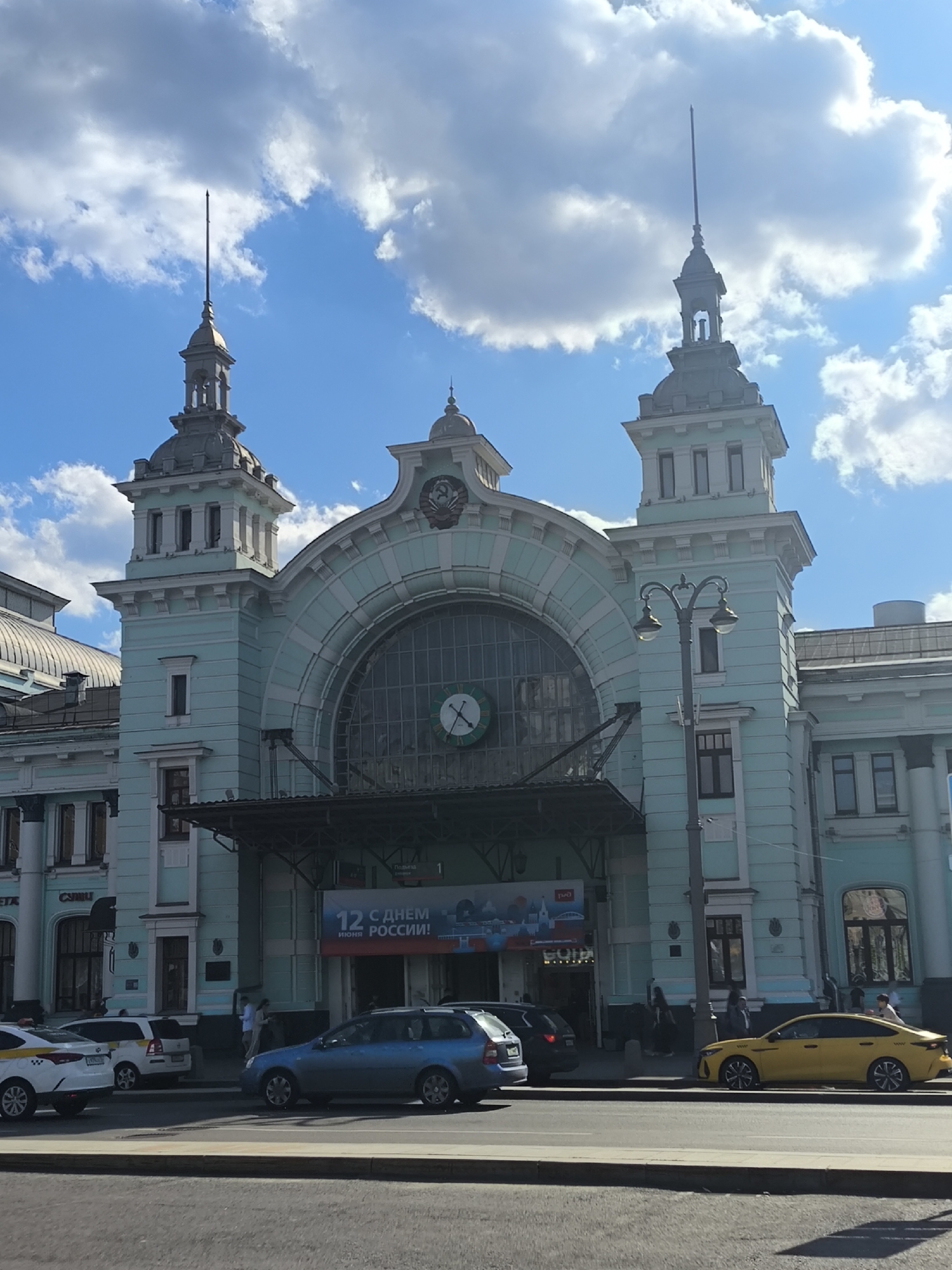 This daytime image captures the grand facade of a historic building, likely a train station, under a bright blue sky dotted with fluffy white clouds. The building is painted a distinctive light teal or mint green with white architectural details, featuring two prominent towers with spires on either side of a large, arched central section. A big clock face is visible within the main arched window, and a smaller clock is positioned above the arch. Above the central arch, an emblem resembling the Soviet hammer and sickle is visible.
In the foreground, a multi-lane street is active with several cars, including a white taxi, a white SUV, a blue sedan/wagon, and a yellow sedan (likely another taxi). A few people are seen walking on the sidewalk near the building's entrance, and a decorative black street lamp stands in front.
A large banner hangs prominently above the entrance, displaying the Russian text "12 С ДНЕМ РОССИИ!" (12 HAPPY RUSSIA DAY!), indicating a celebration of Russia Day, which falls on June 12th. This text, combined with the building's architectural style and the Soviet-era emblem, strongly suggests the location is in Russia, likely a major city like Moscow, and the structure is a significant railway terminal. The scene depicts a sunny day with clear weather, illustrating a typical urban setting with daily activity.