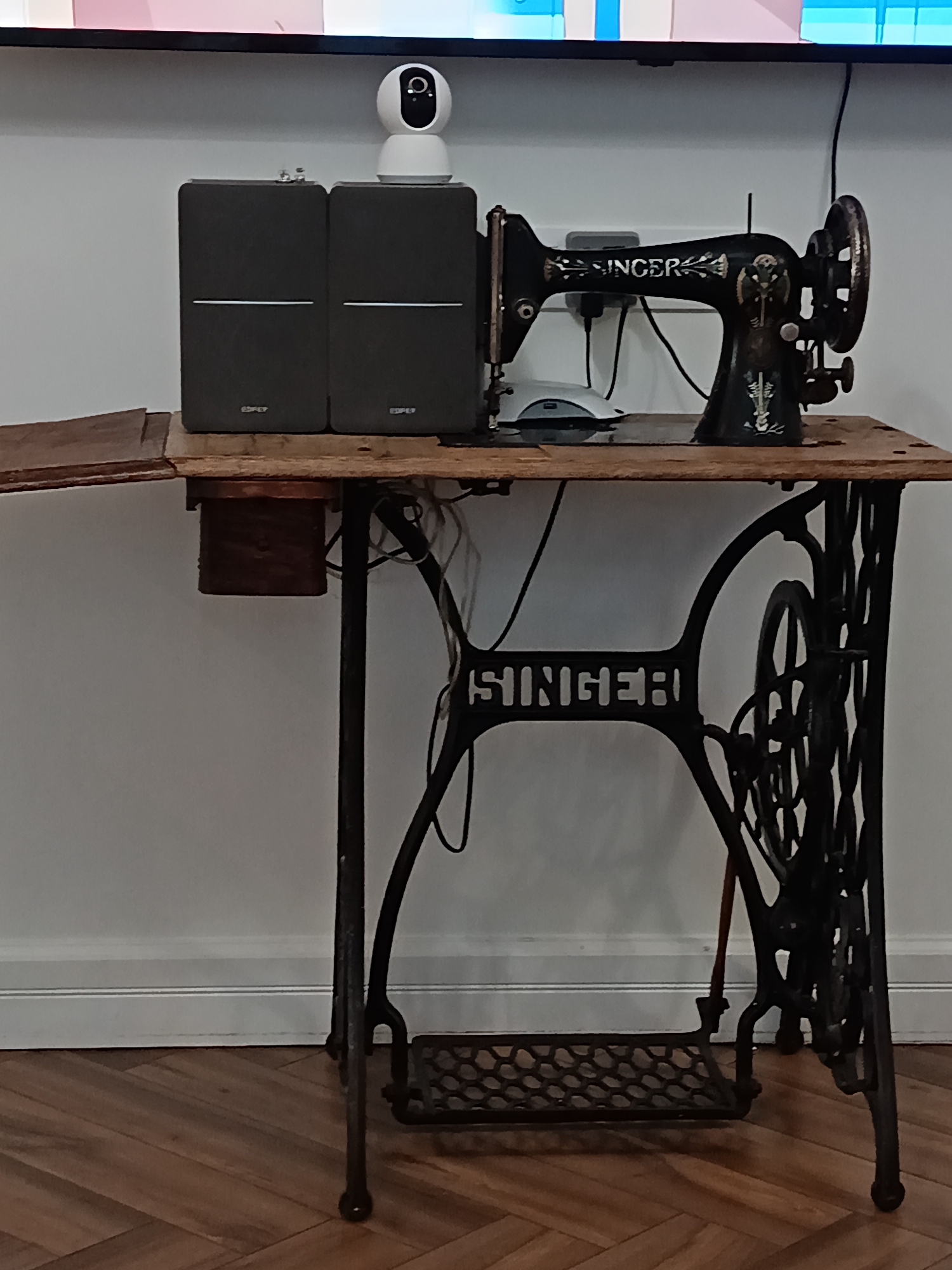 The image shows a vintage Singer sewing machine mounted on a wooden table. The table is supported by an ornate cast-iron base with a foot pedal. On the table, next to the sewing machine, are two dark grey speakers and a white security camera. The scene appears to be indoors, with a white wall and wooden flooring. The sewing machine is black with decorative gold detailing and the "SINGER" logo. The overall setting suggests a domestic environment, possibly a workshop or living space. The location of Pokrovskoe, Russia, is not visually evident in the image.