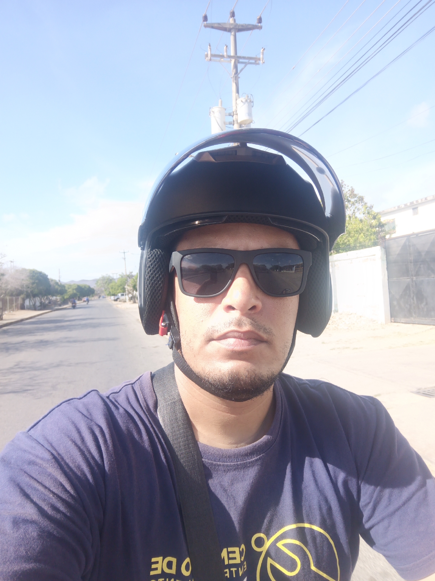 A selfie shot in daylight shows a man wearing a black motorcycle helmet and sunglasses. He has a short beard and is wearing a dark blue t-shirt with a yellow design on the front. A strap is visible across his chest, possibly belonging to the helmet or a bag.
The background shows an asphalt road stretching into the distance, with power lines and a utility pole visible above. A few trees line the road, and buildings are visible on the right. The sky is blue with some clouds.
The location is Juan Griego, Venezuela.