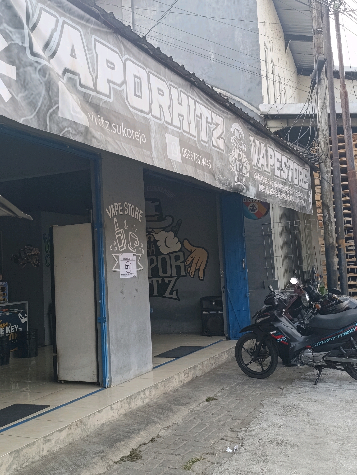 The image shows the storefront of "Vaporhitz Vape Store" located in Karangsono, Indonesia. The store has a large banner with the store's name and contact information, including a phone number and social media handles. Inside the store, there are shelves stocked with vape products. Outside the store, two motorcycles are parked. The building is a single-story structure with a gray facade. The street in front of the store is paved with concrete. There are power lines and a utility pole on the right side of the image. A sign on the store's wall indicates that it is a vape store and has a warning sign stating "PERINGATAN (21+) HANYA UNTUK USIA 21 KE ATAS," which translates to "WARNING (21+) ONLY FOR AGES 21 AND ABOVE."