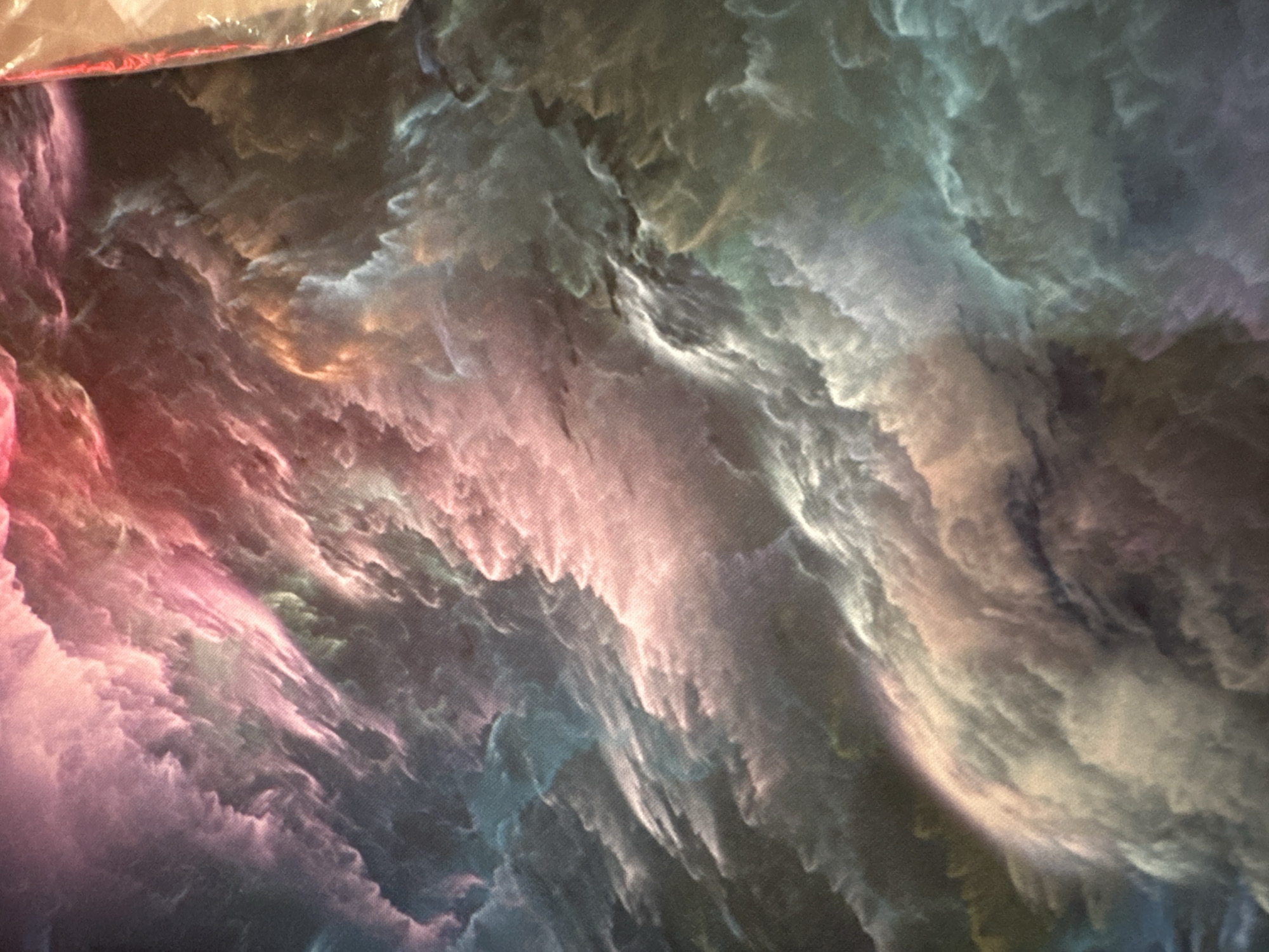 The image shows a close-up, abstract, and textured surface with swirling patterns of color. The colors range from deep blues and greens to vibrant pinks and reds, interspersed with white and gray. The texture appears to be fabric or a similar material, with a visible weave. In the upper right corner, a portion of a transparent plastic bag with a red edge is visible. There are no people, identifiable objects, or environmental context that would place this image in a specific city or location.