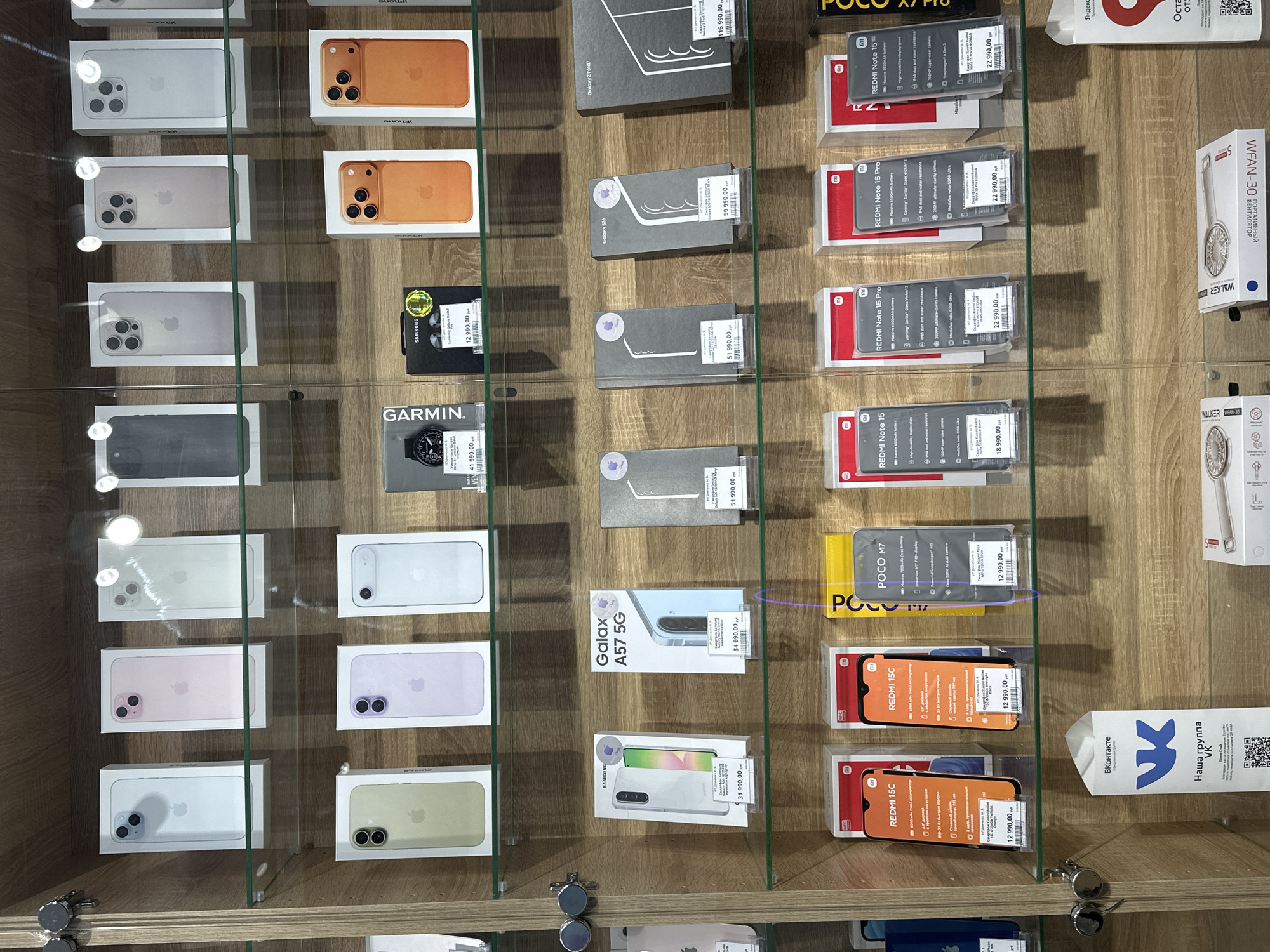 This image depicts a display case in a store, likely located in Chaikovsky, Russia. The case contains various boxed smartphones and accessories. Brands prominently featured include iPhone, Samsung, Redmi, and Poco. Prices are visible on the boxes, indicating they are for sale. Accessories include smartwatches (Garmin), portable fans (Walker), and other phone models. The boxes are neatly arranged on wooden shelves within the glass-fronted display case. The store is affiliated with VKontakte, a social media platform.