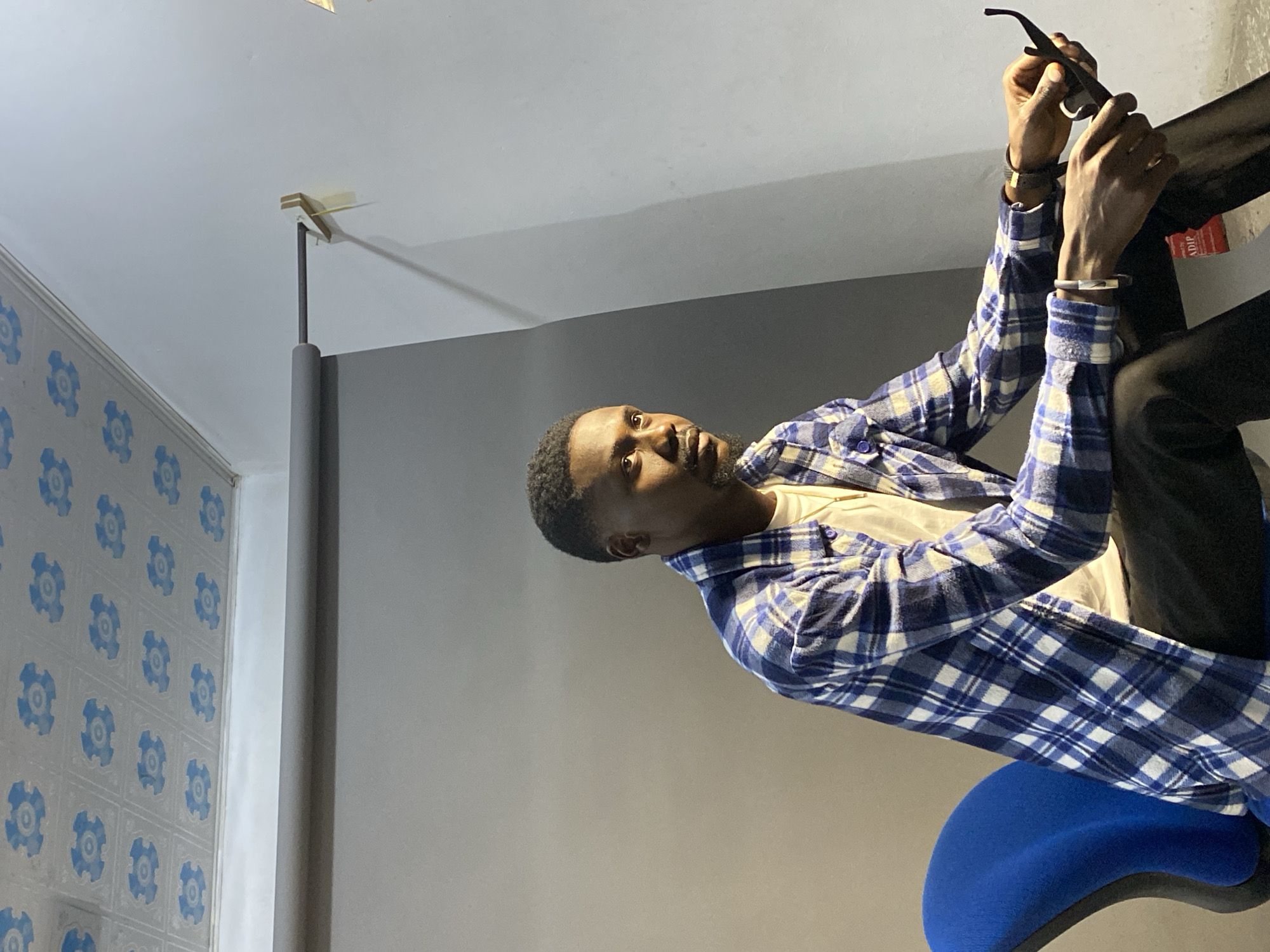 A man sits on a blue chair, facing right. He's wearing a blue and white checkered shirt, a beige undershirt, and black pants. He is holding a pair of sunglasses with both hands. The background consists of a grey wall and part of a wall decorated with blue floral patterns. A vertical support beam is also visible in the scene. The image appears to be indoors, although specific details about the location in Jos, Nigeria, are not present in the image.