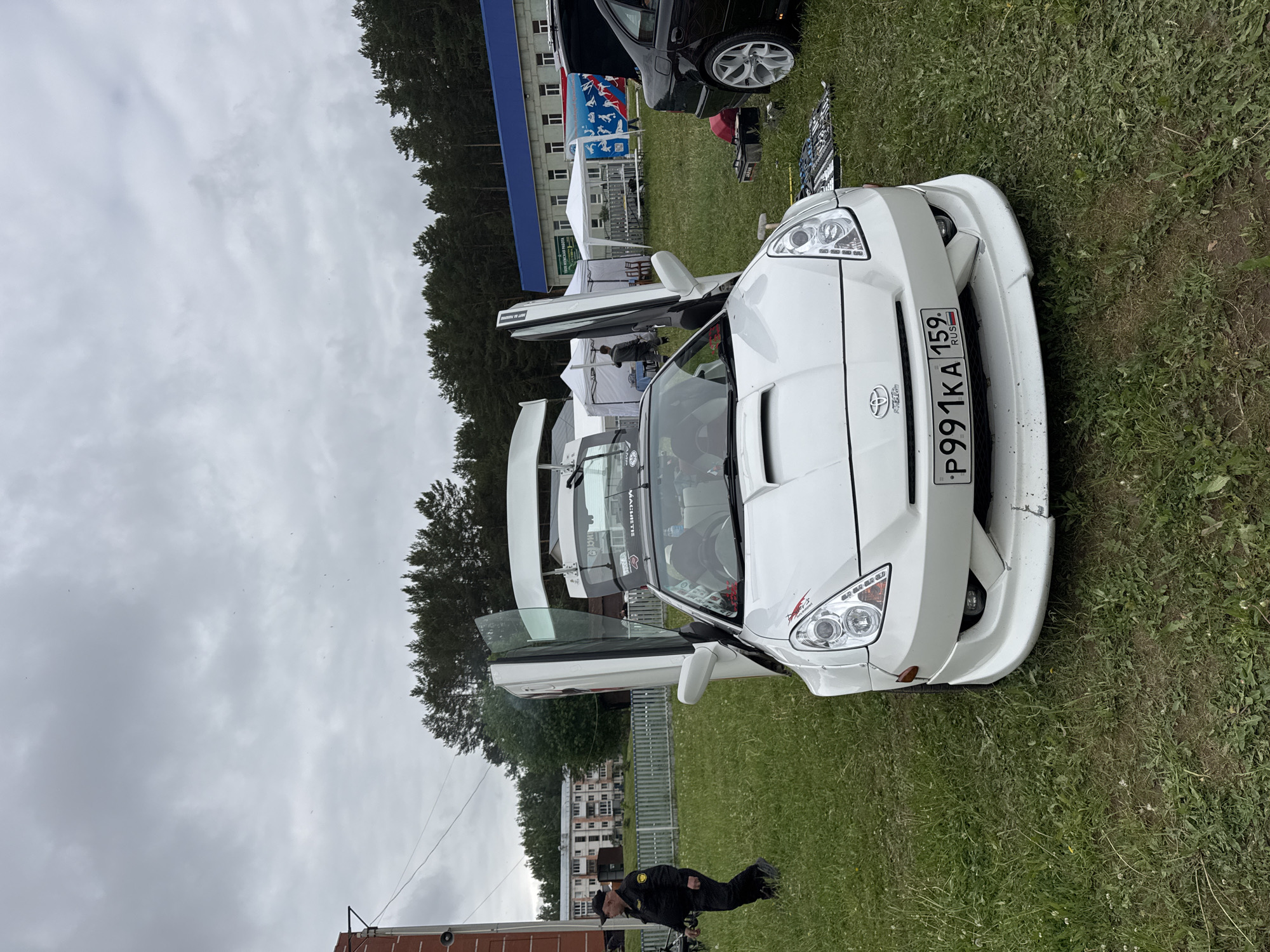 The image shows a white Toyota Celica with its doors open, parked on a grassy field. The license plate reads "P991KA 159 RUS." The car appears to be modified with a custom body kit and aftermarket wheels. In the background, there are trees, a building with a blue roof, and other cars. A person dressed in black uniform is next to a small trailer on the left of the image. The location is most likely in or near Chaikovsky, Russia, as indicated by the license plate. The sky is overcast, suggesting a cloudy day.