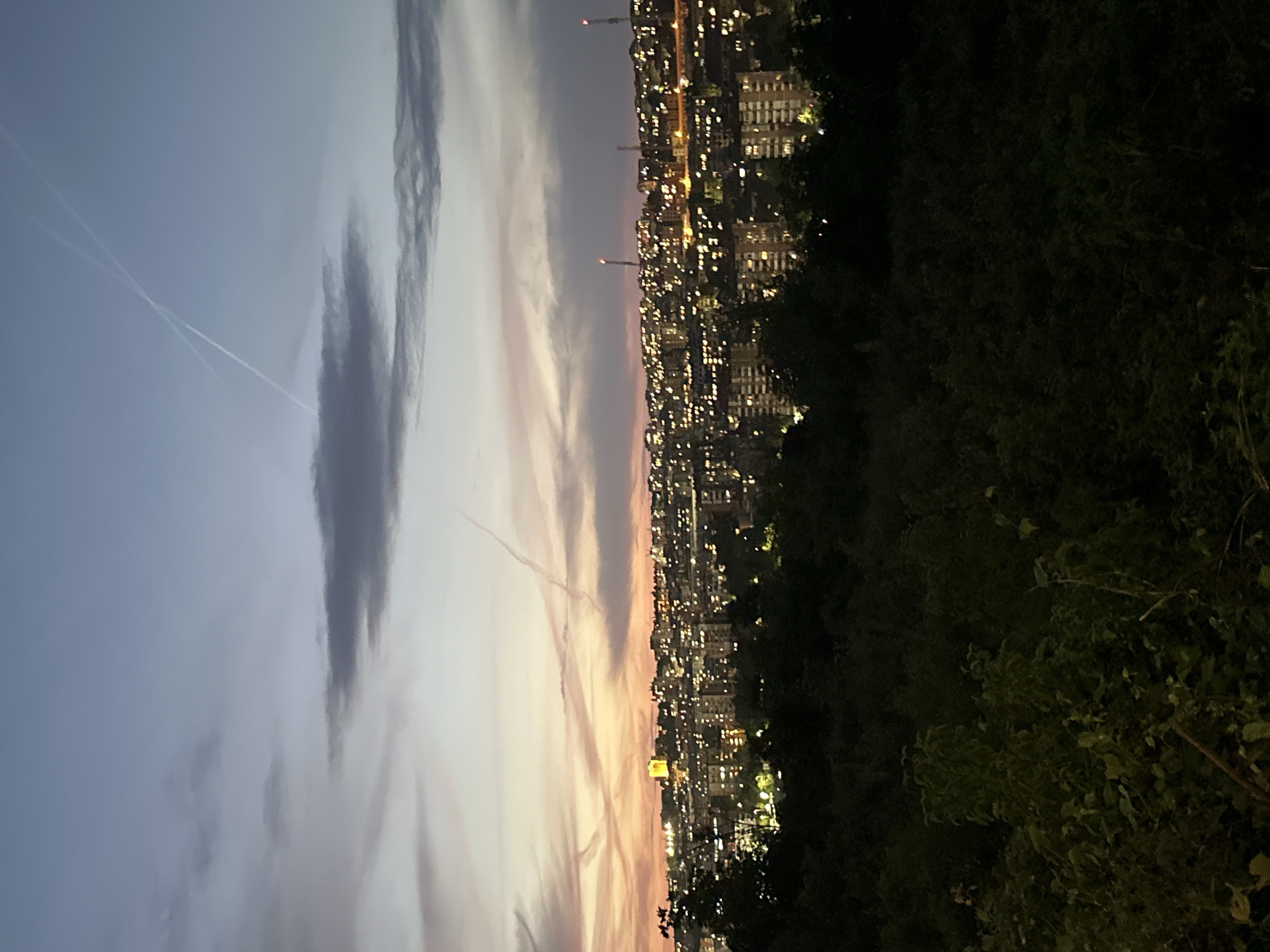 Here's a description of the image:
**Scene:** An elevated, slightly skewed view overlooking a city at dusk or twilight.
**Environment:** The sky transitions from a pale blue-grey at the top to shades of pink and orange towards the horizon. Streaks of clouds are visible, along with contrails from aircraft. The city is densely populated, with buildings illuminated by numerous lights. A dark, indistinct area with trees and foliage is located in the foreground to the right, partially obscuring the view.
**Objects:** Buildings of varying heights, with many windows lit. Two tall radio transmission towers are visible. Contrails are present in the sky. Trees and shrubbery are located in the foreground.
**Location:** It appears to be Veliko Tarnovo, Bulgaria, though this cannot be confirmed definitively from the image alone.