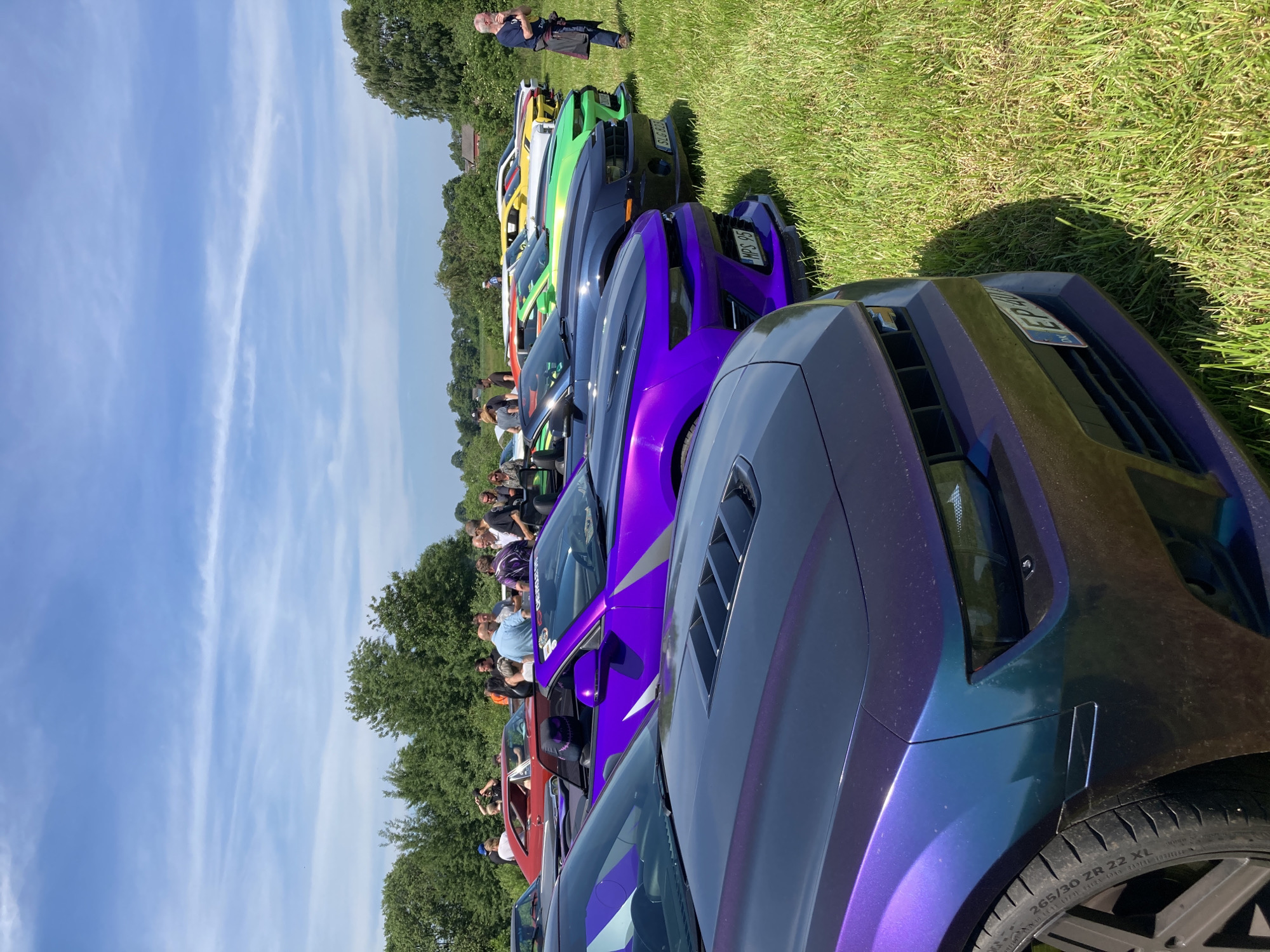 The image depicts an outdoor car meet event in Edenberga, Sweden, under a bright blue sky with scattered clouds. A line of brightly colored Chevrolet Camaros are parked on a grassy field. In the foreground, the hood and front bumper of a dark purple Camaro are visible. Behind it, a vibrant purple convertible Camaro is parked, with a skull-themed pillow visible in the driver's seat. Further down the line, Camaros in bright green, yellow, and white are also displayed. A group of people are gathered around the cars, some standing and talking, others appearing to take photos. A man in a blue shirt and jeans is standing near the center of the gathering, while another man in a dark shirt and jeans is walking away from the cars on the right side of the frame. The background features a line of trees and some scattered buildings, suggesting a rural or semi-rural setting. The overall atmosphere appears to be relaxed and social, typical of a car enthusiast gathering.