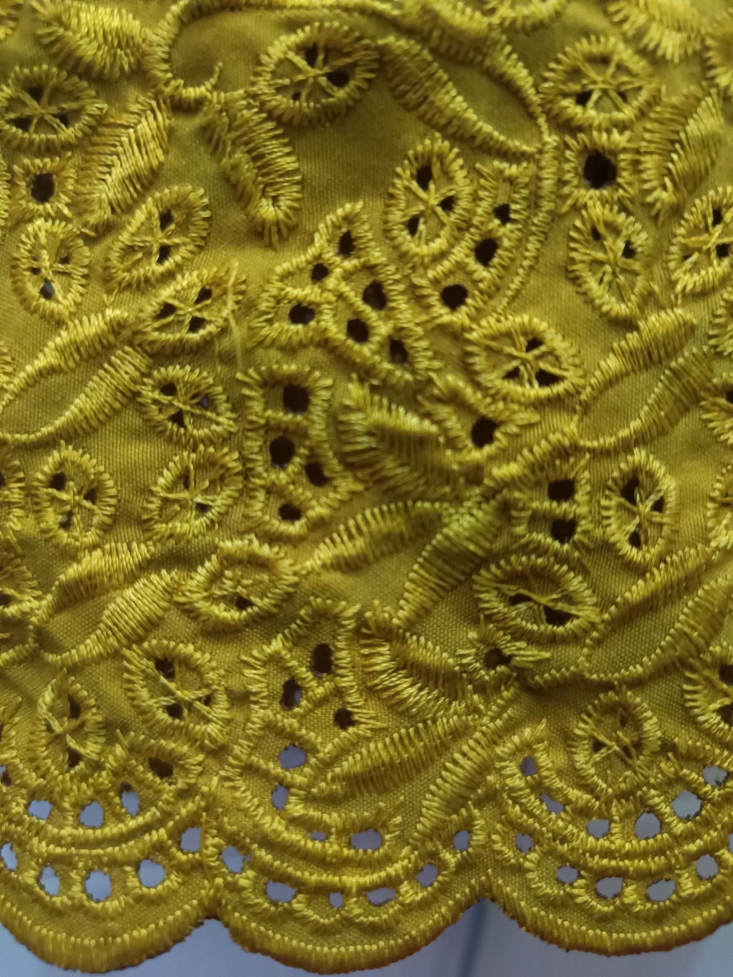 A close-up, high-angle shot shows a piece of embroidered yellow fabric. The embroidery features intricate floral and circular patterns with cut-out details, creating a delicate, lacy effect. The fabric appears to be a cotton or linen blend, with a slightly textured surface. The lighting is even, with no harsh shadows, highlighting the detail and texture of the embroidery. The background is out of focus and appears to be a light neutral color, possibly a wall or another piece of fabric. The specific location in Gunung Sekar, Indonesia, is not determinable from the visual information provided in the image.