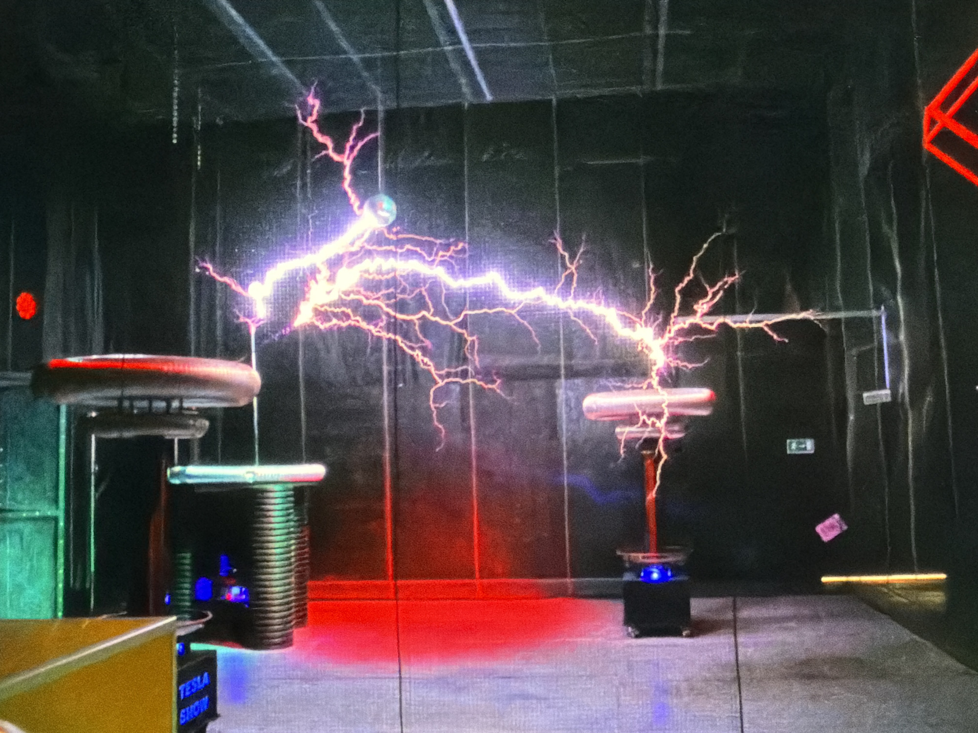 The image depicts a Tesla coil demonstration in a dark room, likely an exhibition or show. Two coils are visible, one on the left and one on the right, with electrical arcs discharging between them. The arcs are pink/purple and branch out like lightning. The coils consist of circular toroidal electrodes on top of cylindrical structures. The platform under the left coil has "TESLA SHOW" printed on its side. The background is a dark wall, possibly concrete or painted. There is a red light shining on the wall behind the left coil. In the background on the right, there is a green exit sign. A small object in a pink wrapper appears to be floating near the exit sign. Part of a red geometric structure is visible in the upper right corner. The floor is concrete. The location is unknown, as is the city.