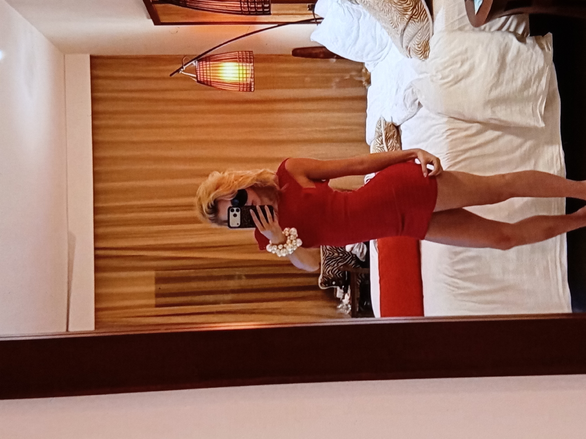 A woman in a red dress is taking a mirror selfie in a room with a bed, a lamp, and a wooden-slatted wall. The location cannot be determined to be Yerevan, Armenia without additional information.