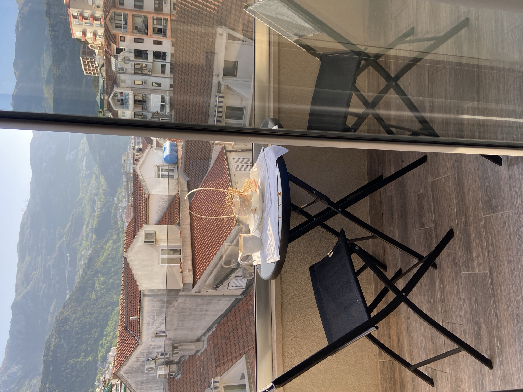 The image captures a serene outdoor scene from a balcony overlooking a town and majestic mountains in Budva, Montenegro. The balcony features a light wooden plank floor and a transparent glass railing that provides an unobstructed view.
On the balcony, a small, round table with a dark top is set with a white ceramic mug, a folded white cloth or napkin, and a small, light-colored woven basket, suggesting a moment of quiet enjoyment like coffee or breakfast. Two black folding chairs with black fabric seats and metal frames are positioned around the table, ready for use. The clear reflection of one of the chairs and its structure is visible on the glass railing.
Beyond the balcony, the view opens up to a densely packed town built into a hillside. Numerous buildings with distinctive red tile roofs are visible, interspersed with structures featuring various window styles and balconies. In the background, large, verdant mountains rise steeply, covered in trees and vegetation, stretching into the hazy distance under a clear, bright sky. The lighting indicates it is daytime, likely morning or early afternoon, with clear weather. No people are visible, and there is no discernible text in the image.