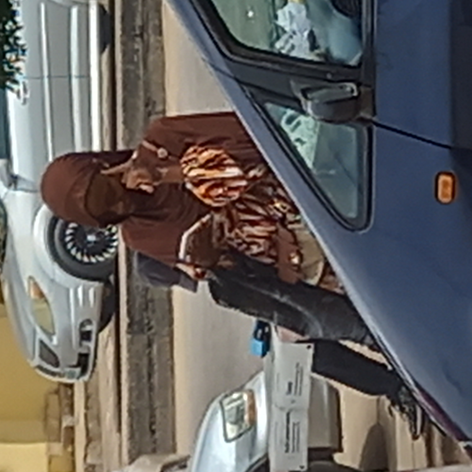 A person with a brown hijab is seen holding a child with a tiger-striped hat while standing next to a dark blue car, possibly in Dadi Riba, Nigeria. A silver car is visible in the background. The person is wearing black pants and shoes. There appears to be a light-colored object or structure partially visible in front of the blue car.