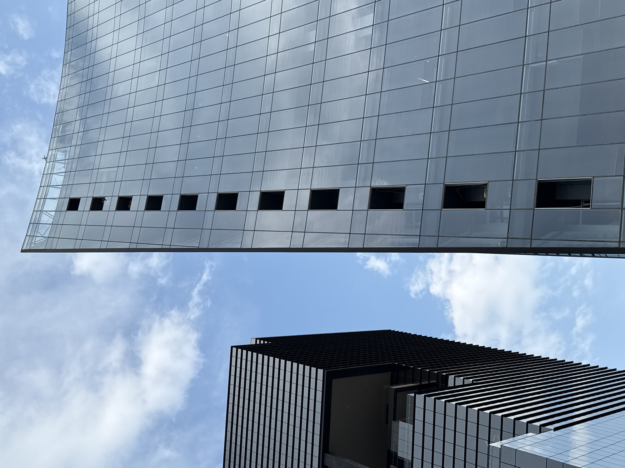 The image shows two buildings from a low-angle perspective against a sky with scattered clouds. The building above has a glass facade with regularly spaced square windows near its lower edge, some of which appear to be open or damaged. The building below is darker, with a layered, striated design along its top, potentially an architectural feature. The angle suggests the photo was taken in a dense urban area.
The location is Puebla, Mexico.