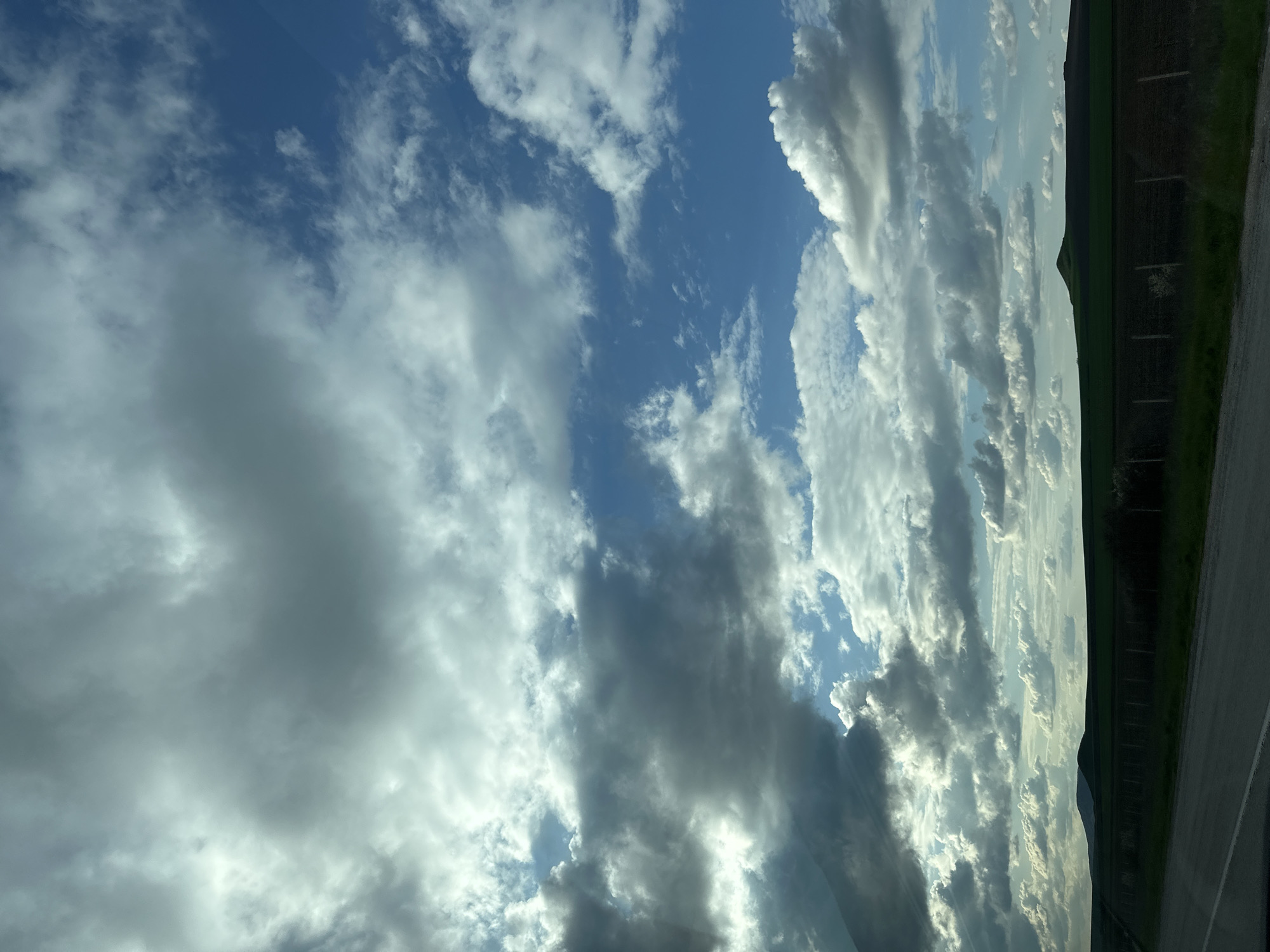 The image depicts a cloudy sky with visible patches of blue. The clouds are varied, ranging from fluffy white to dark grey storm clouds. In the upper right corner, there is a glimpse of land, likely a field and possibly some trees. A grey surface, probably a road, is visible at the very bottom right. The image is rotated 90 degrees counterclockwise. The location, Zlatina, Bulgaria, is not discernible from the visual content of the image.
