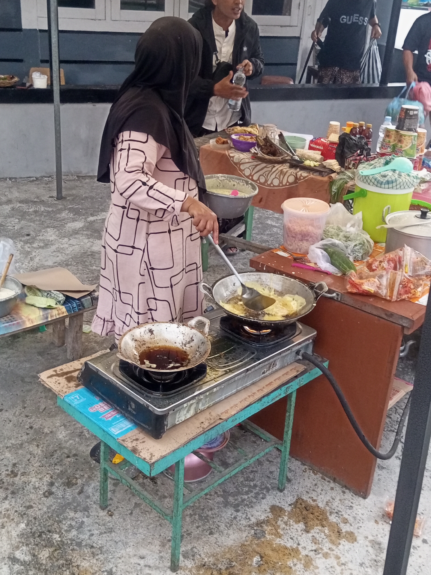 In Pandansari, Indonesia, a woman wearing a black hijab and a pink patterned dress is cooking outdoors at a street stall. She is actively frying food in a wok placed on a portable gas stove. The stove is positioned on a light blue and green metal table. Behind her, a stall displays various food items, drinks, and other goods, including bottles of sauce, cans, and packaged snacks. Several people are visible in the background, some standing and interacting, indicating a busy market or food street environment. The ground surface is concrete, with some spilled food or dirt scattered around the cooking area. The overall scene depicts a typical street food vendor operation in a bustling Indonesian city.