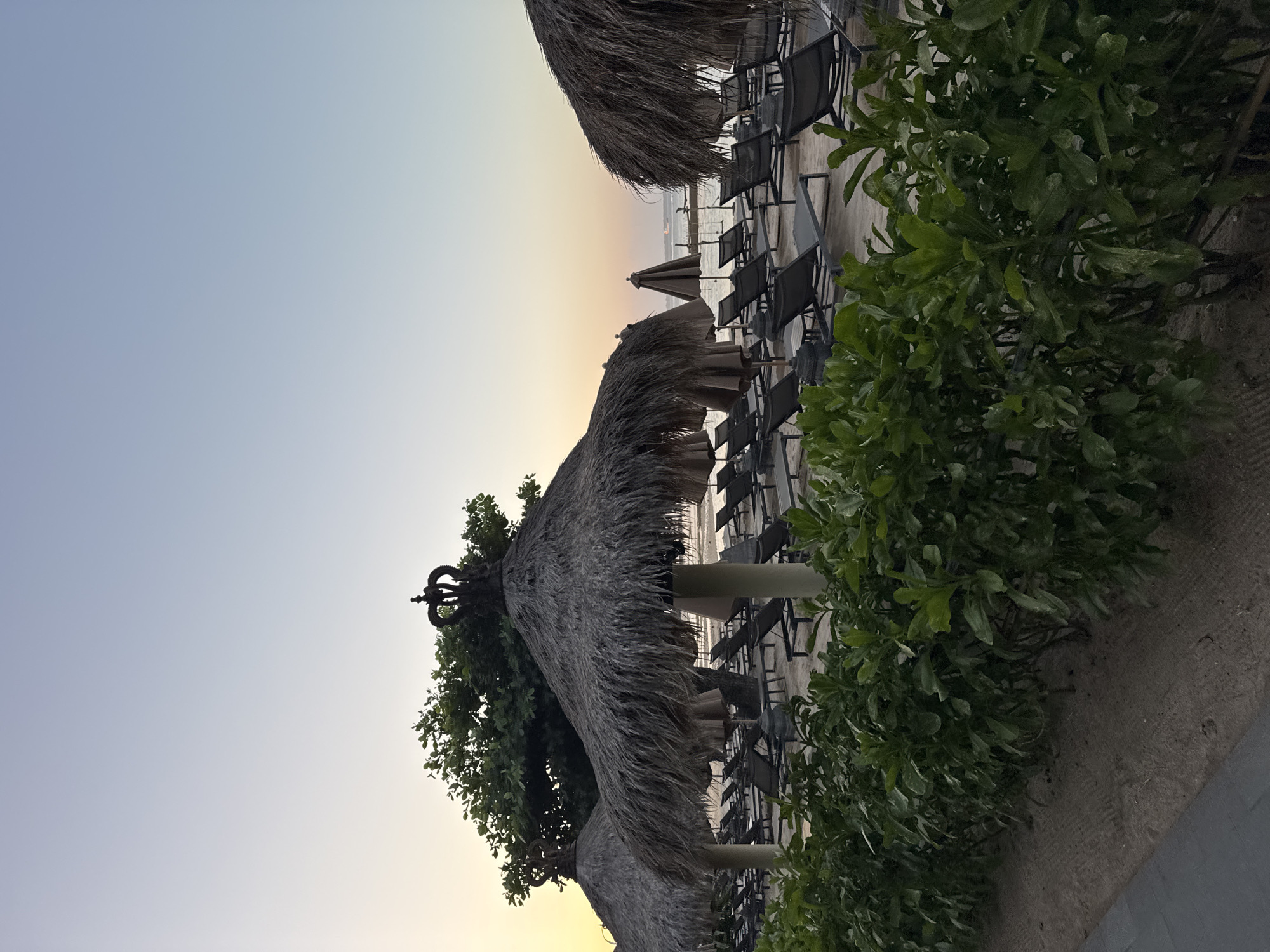 The image depicts a tranquil beach scene at dusk in Kuta Selatan, Indonesia. Several thatched umbrellas with decorative crown finials stand on the sand, casting shadows over rows of empty lounge chairs. Lush green foliage lines the foreground, separating the lounge area from a paved walkway. In the background, the calm sea stretches to the horizon under a soft, gradient sky transitioning from a pale yellow at the horizon to a light blue overhead. The overall atmosphere is peaceful and serene, suggesting a resort setting preparing for the evening.