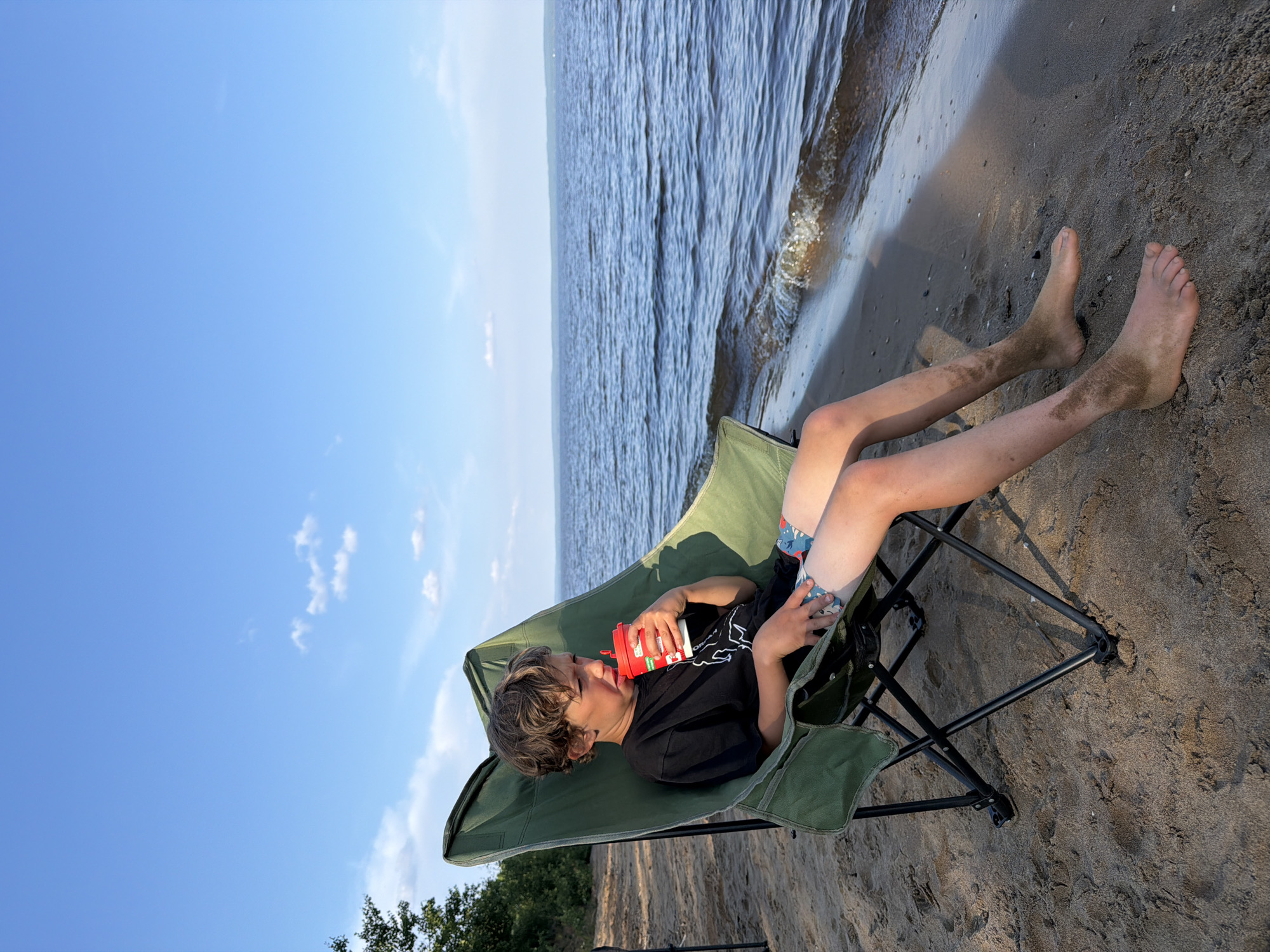 A young Caucasian boy with brown, tousled hair sits in a green, folding camping chair on a sandy beach. He is wearing a black t-shirt with a white graphic and patterned blue swim trunks. The boy is holding a red and white cup with a straw and appears to be drinking from it. His legs are dirty with sand, and his feet are bare and also covered in sand. The chair is positioned facing a large body of water, likely a lake or river, with a distant shoreline visible under a partly cloudy blue sky. The scene takes place outdoors on a sunny day. The location is the Kama River beach in Chaikovsky, Russia.