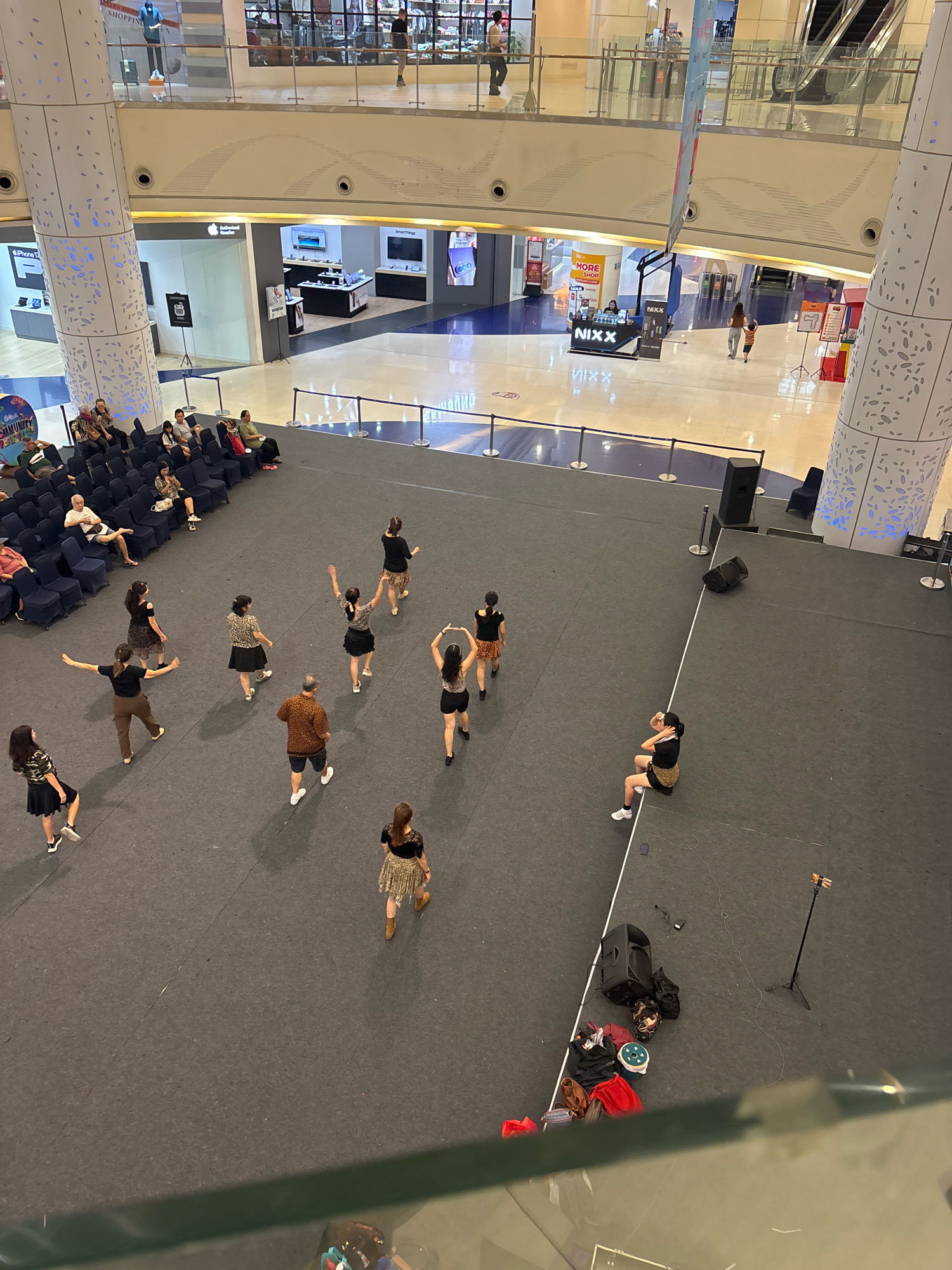 This image captures an aerial view of a dance rehearsal taking place on a dark carpeted floor in what appears to be a public atrium or shopping mall, likely in North Jakarta, Indonesia, given the branding and architectural style. Several individuals, predominantly women, are engaged in various dance poses, some in unison and others individually. They are dressed in casual attire, with many wearing skirts and tops. Spectators are seated on dark chairs arranged in rows on the left side of the frame, observing the performance. The environment is spacious and modern, with elevated walkways and storefronts visible in the background. Audio equipment, including speakers and a microphone stand, are present on the dance floor, indicating a performance or practice session. The lighting suggests an indoor setting, likely during daytime hours.