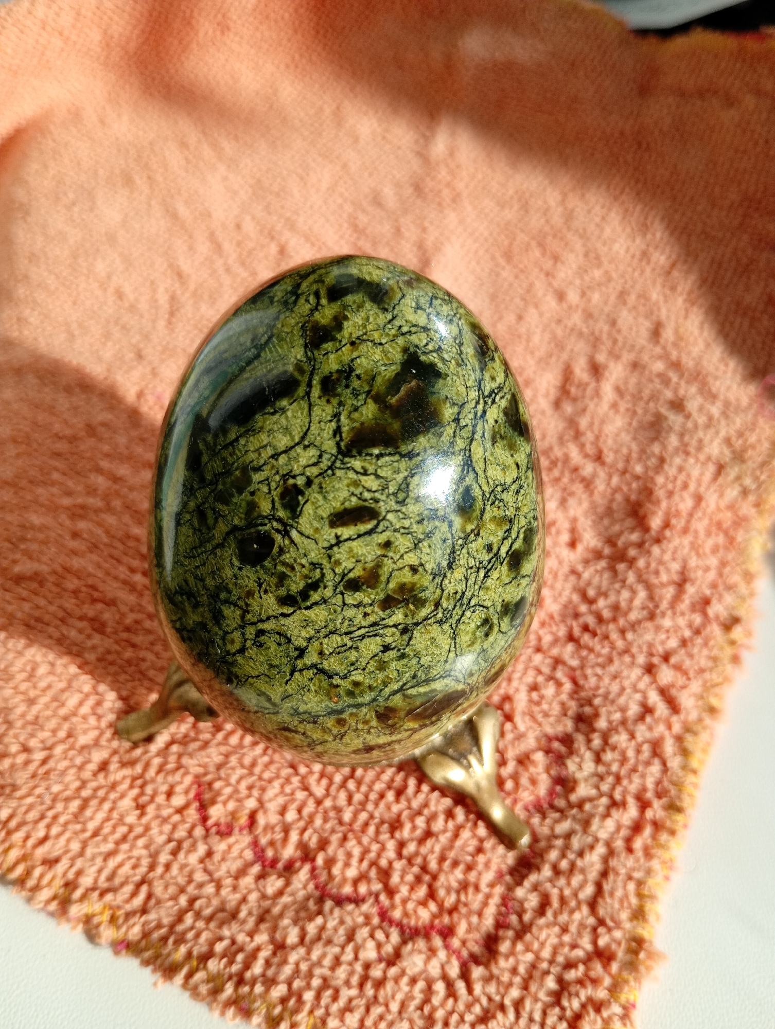 A polished, egg-shaped stone, possibly jade or serpentine, rests on a small brass stand with three claw-like feet. The stone displays a variegated green and dark brown pattern with intricate black veining. It is positioned on an orange, terrycloth-like fabric with a textured surface. A shadow is cast across the fabric from the upper left. The scene is illuminated by bright sunlight, creating highlights on the stone and casting a distinct shadow. The location is the city of Avanhard, Ukraine.