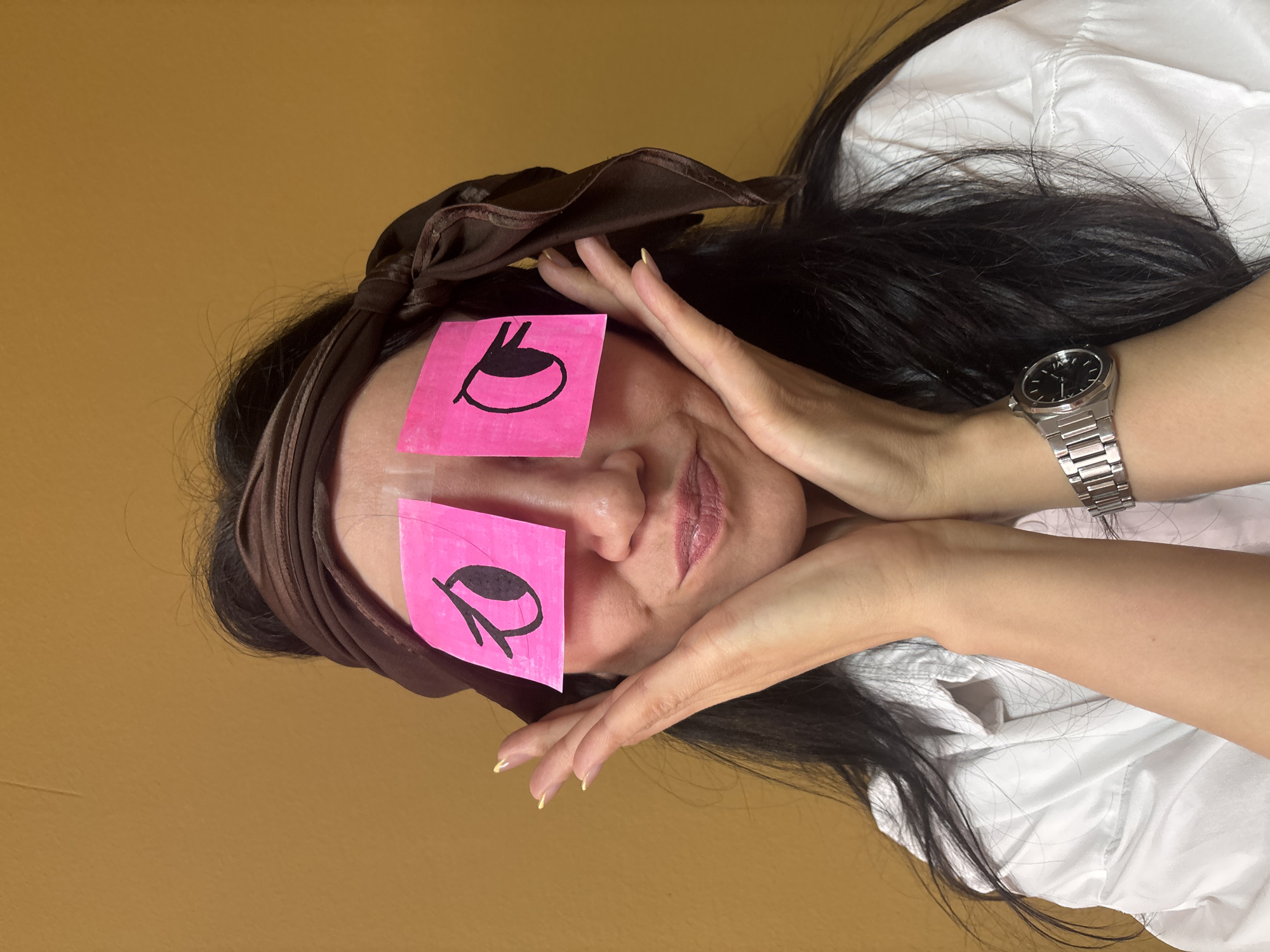 A person with long, dark hair and a brown satin-like headband is depicted lying down against a plain mustard-yellow background. Two bright pink sticky notes are placed over their closed eyes, each featuring a large black outline of an open eye drawn on it. The person's hands are gently positioned around their face, framing it. A silver-toned wristwatch with a black face and white hands is visible on their left wrist. Their lips are slightly parted, and light-colored nail polish is visible on their fingers. They are wearing a white collared shirt. The image, taken from an overhead perspective and rotated on its side, suggests a moment of rest, sleep, or playful fatigue in an indoor setting in Chaikovsky, Russia. There is no visible text, only the drawn eye symbols on the sticky notes.