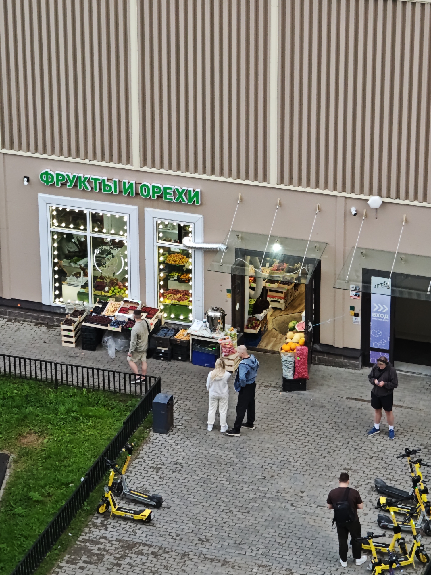 The image depicts an outdoor fruit and nut stand in front of a building with a textured facade. A prominent green sign above the entrance reads "ФРУКТЫ И ОРЕХИ" (Fruits and Nuts). The stand displays various fruits and vegetables in crates and on tables, with some items visible through the storefront windows. Several people are present: a man in a light-colored shirt and shorts stands near the fruit display, looking at his phone; a couple, a woman in white and a man in a blue jacket, are standing near the entrance; and another man in a dark jacket and shorts is also looking at his phone. A group of yellow electric scooters are parked on the paved area in the foreground, with a black trash can nearby. The environment suggests an urban setting, likely a commercial street or marketplace. The image appears to be taken from an elevated perspective.