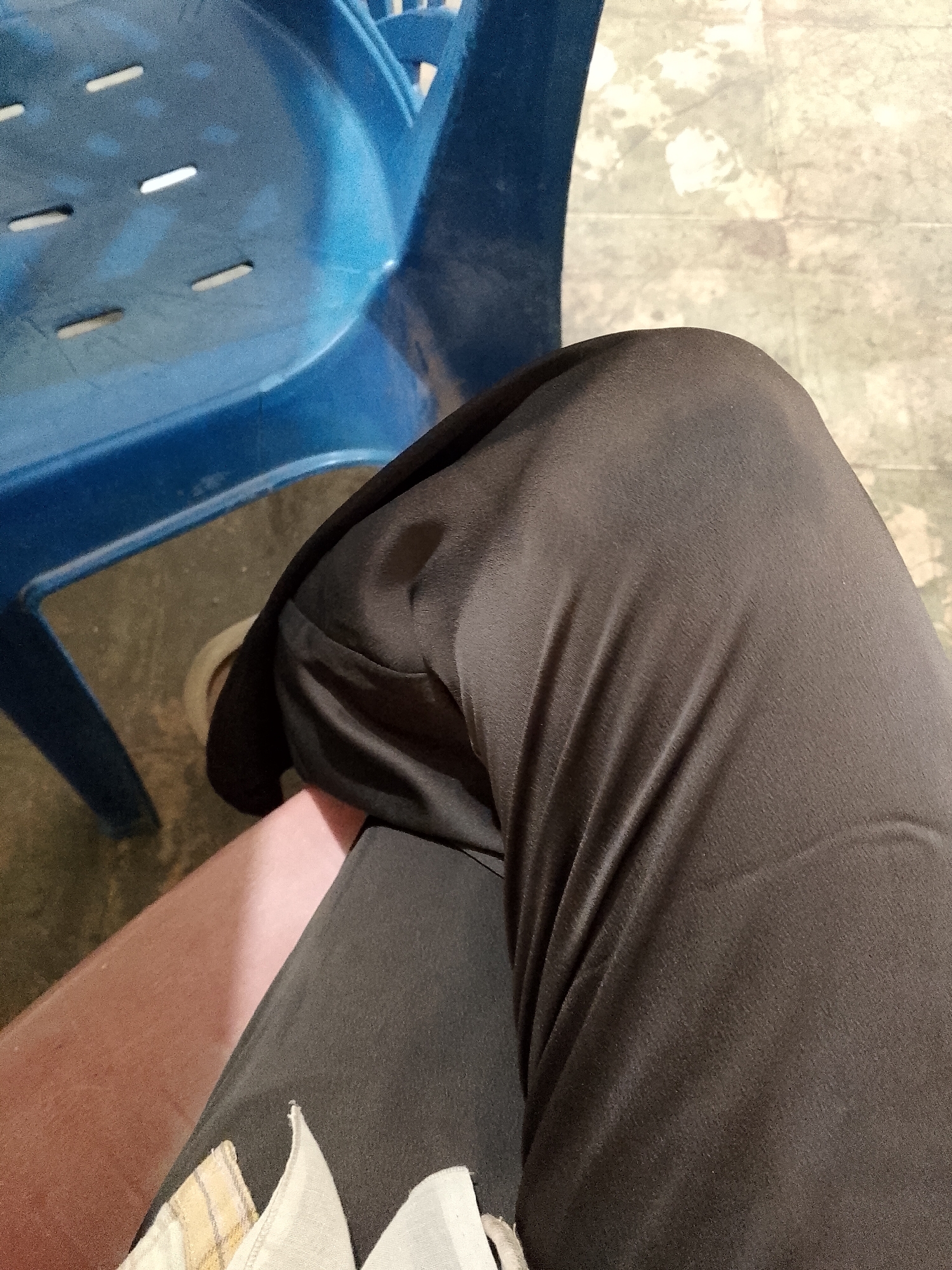 The image shows a close-up view of a person's legs and a portion of a blue plastic chair. The person is wearing dark-colored pants that are wrinkled, and a portion of their thigh is visible with a light-colored garment underneath. The background consists of a patterned floor and the blue chair, which has a slatted design. The scene appears to be indoors or in a sheltered outdoor area. The image does not contain any specific details that would allow for identification of its location within Lhokseumawe, Indonesia.